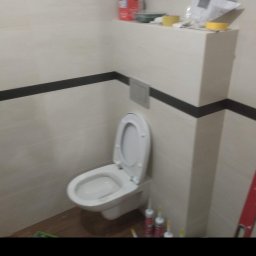 JARDAR SPÓŁKA Z OGRANICZONĄ ODPOWIEDZIALNOŚCIĄ - Zamontowana toaleta w łazience z białymi płytkami i czarnym pasem dekoracyjnym, widoczne silikony uszczelniające i narzędzia na półce.