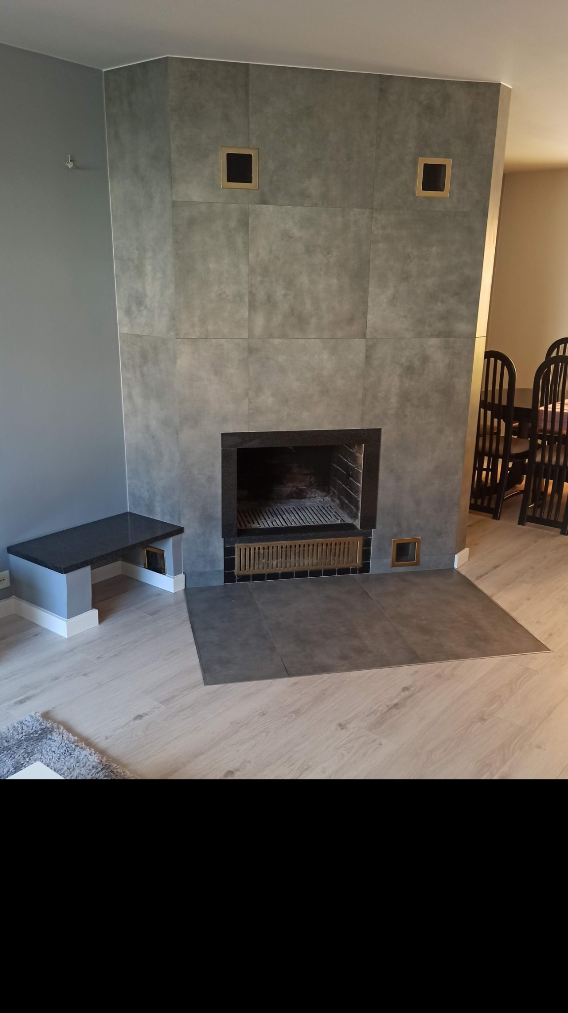 Obudowa kominka z szarych, wielkoformatowych płyt, z czarnym wkładem i złotymi akcentami wentylacyjnymi, uzupełniona o siedzisko z czarnego kamienia.