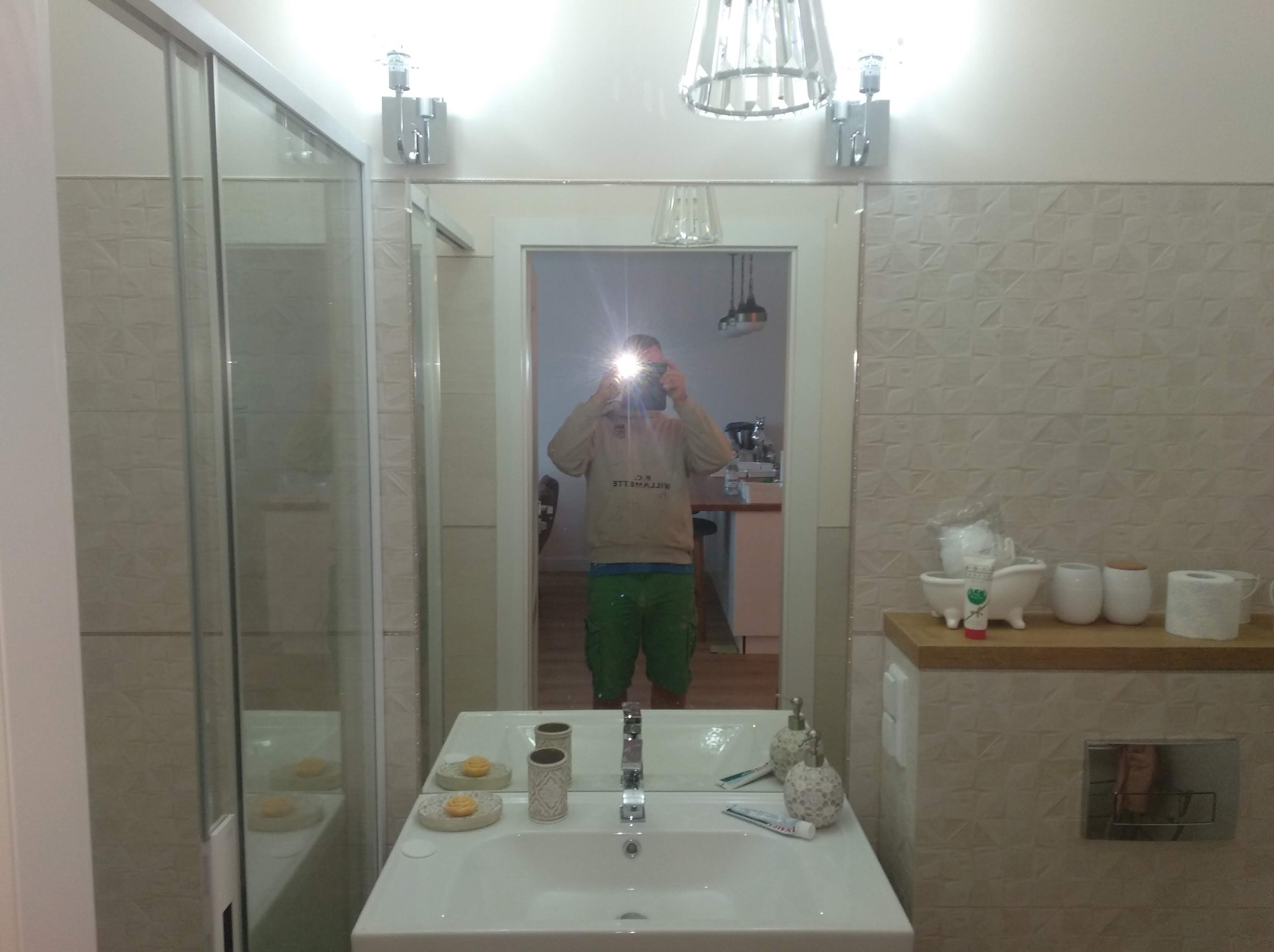 Nowoczesna łazienka z białą umywalką, błyszczącą baterią i dekoracyjnymi akcesoriami, widok z kabiny prysznicowej, odbicie fotografa w lustrze.