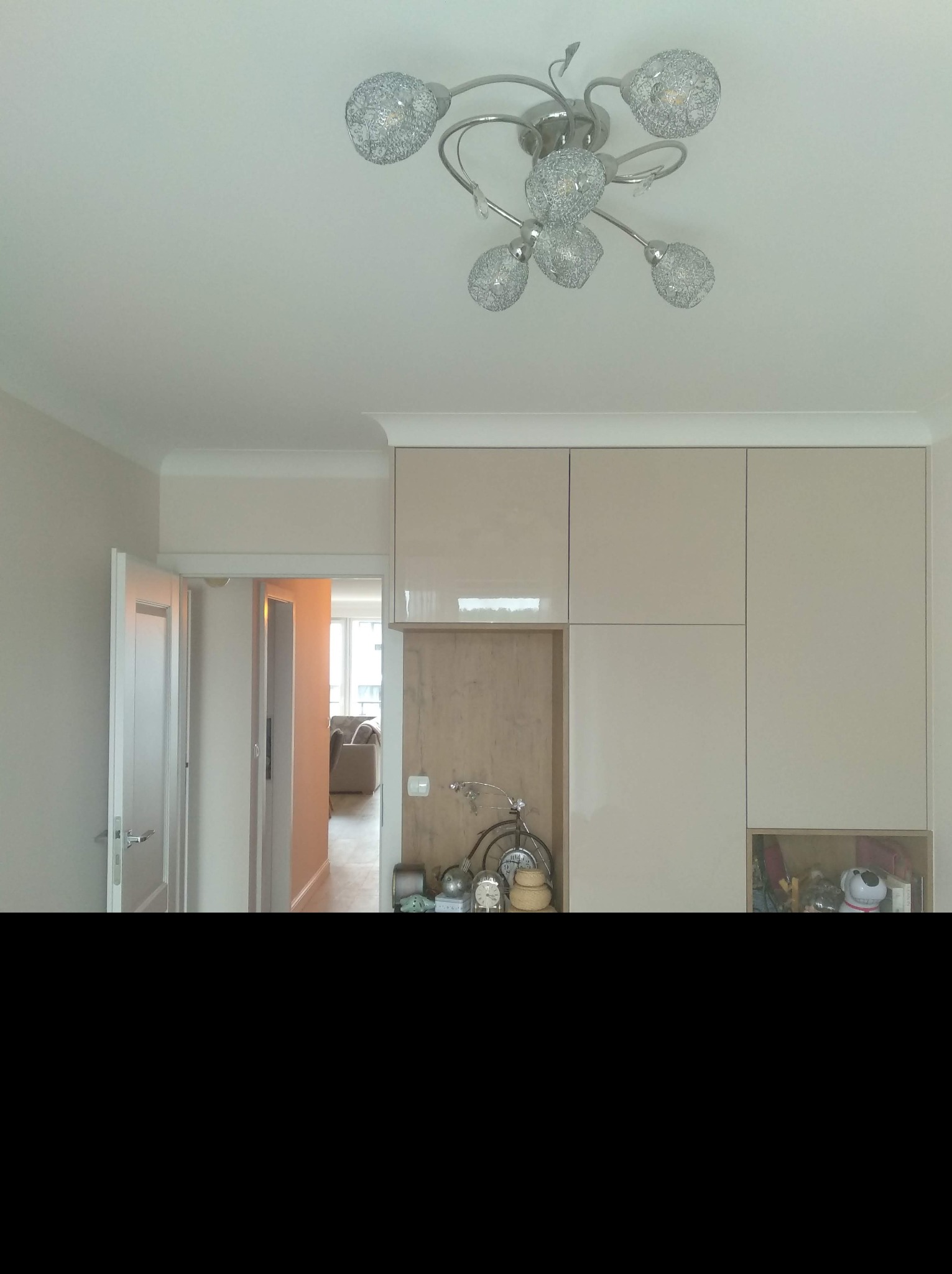 Nowoczesne wnętrze: błyszczące, beżowe szafy w zabudowie pod sufit, dekoracyjna lampa sufitowa z ażurowymi kloszami, otwarte drzwi do pokoju z kanapą w tle.