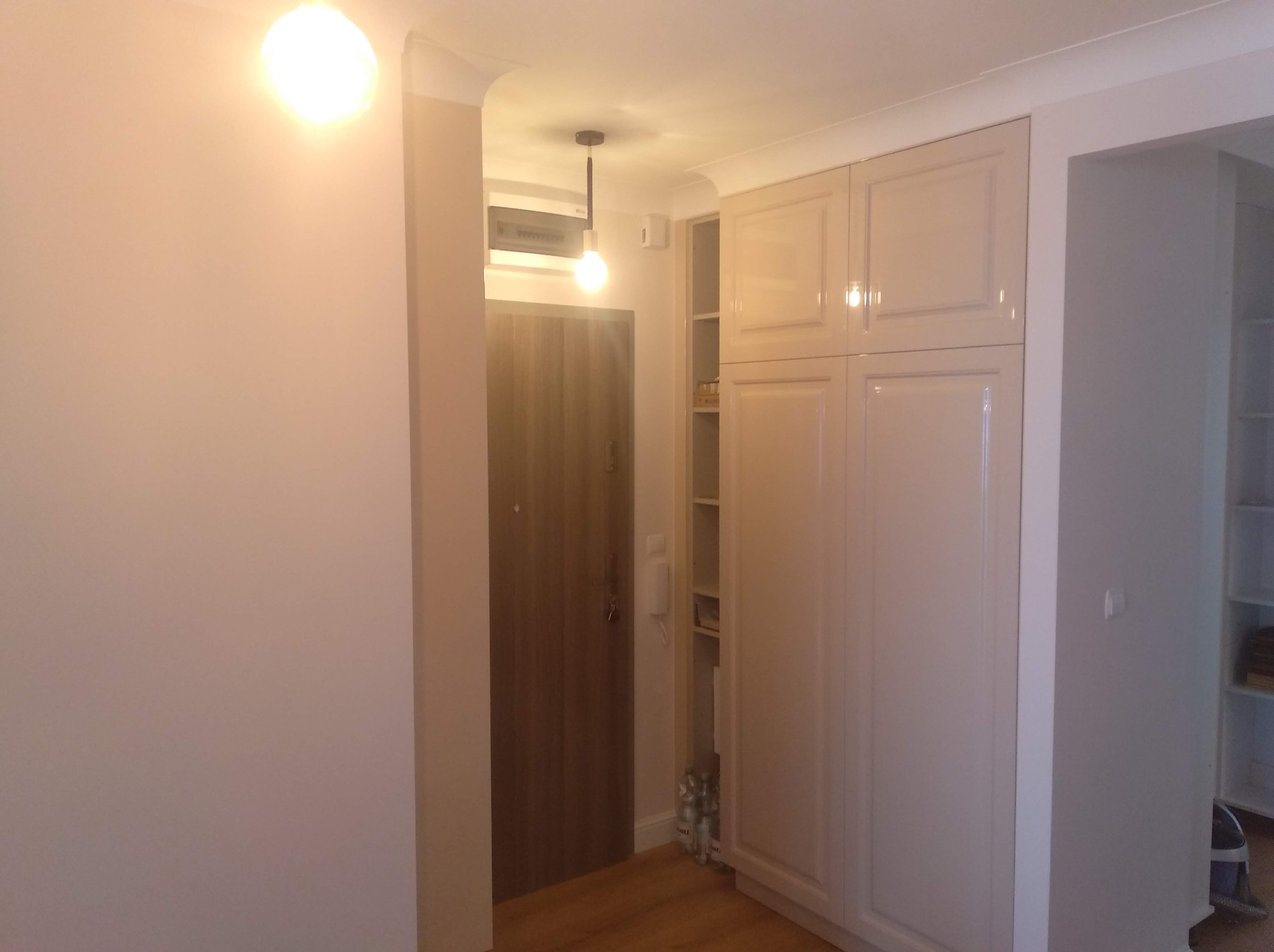 Wykończone wnętrze mieszkania z zabudową szafy w kolorze kremowym i drzwiami wejściowymi z ciemnego drewna, oświetlone wiszącą lampą z żarówką Edison.