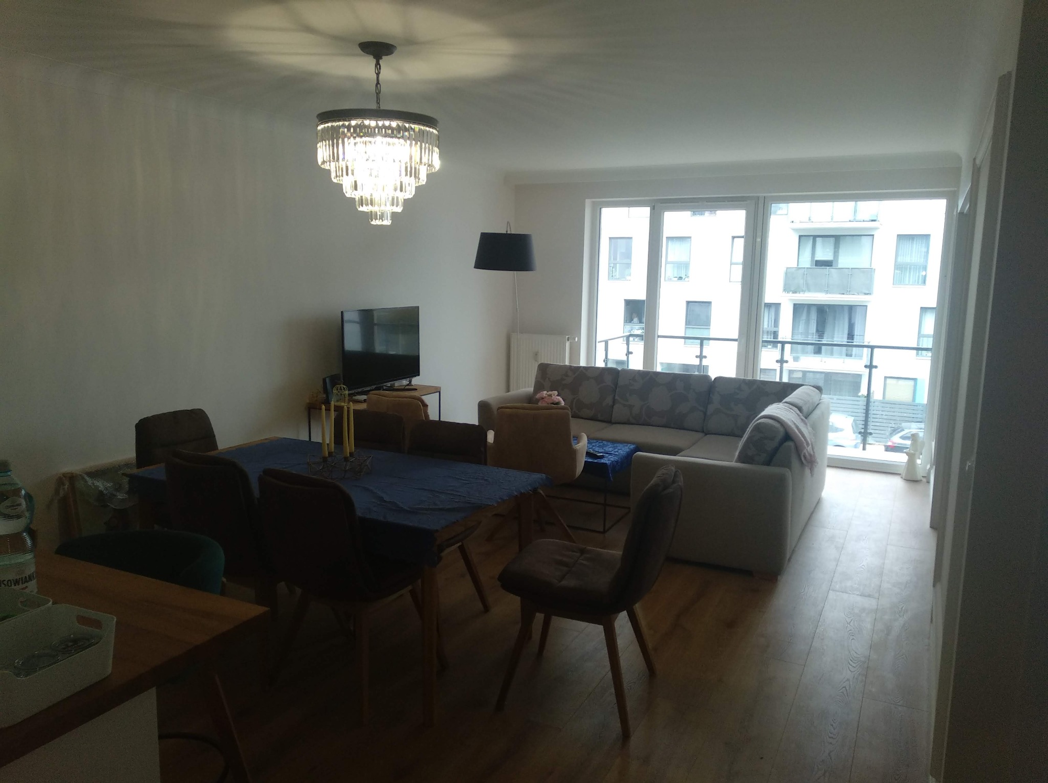 Jasne, przestronne wnętrze mieszkania po remoncie: parkiet, gładkie ściany, kryształowy żyrandol nad stołem z niebieskim obrusem, sofa z widokiem na balkon.