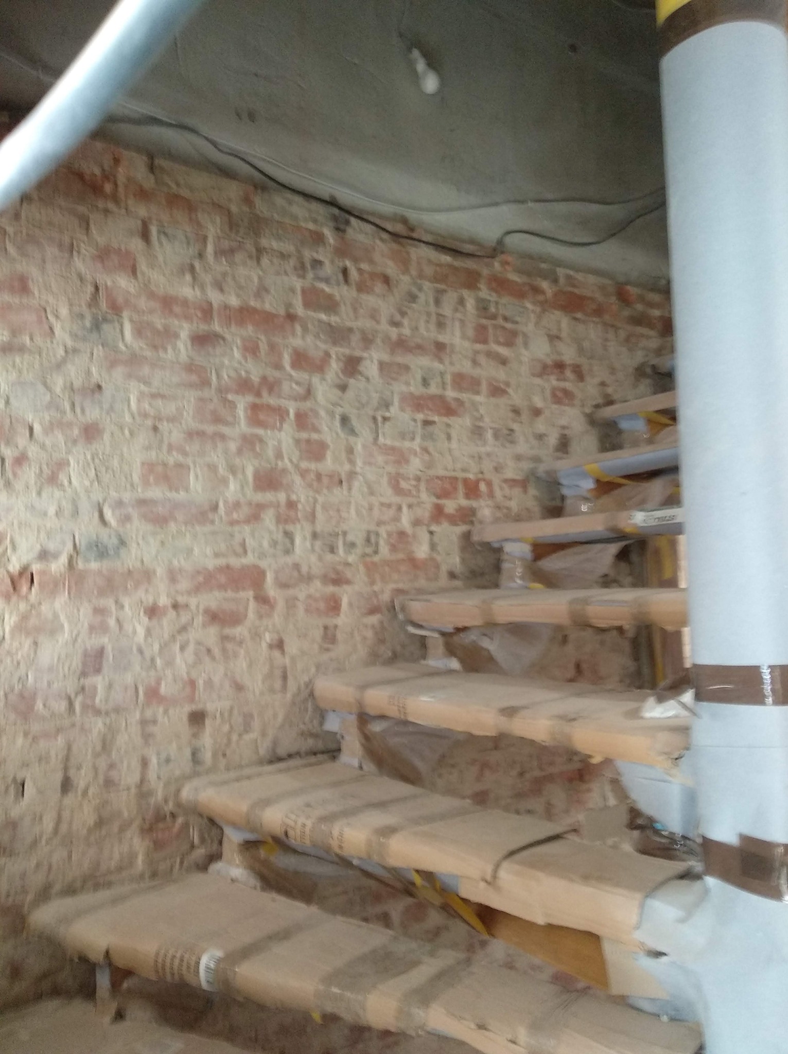 Surowe cegły ściany w trakcie prac remontowych, schody tymczasowo zabezpieczone kartonem, widoczna instalacja elektryczna pod sufitem.