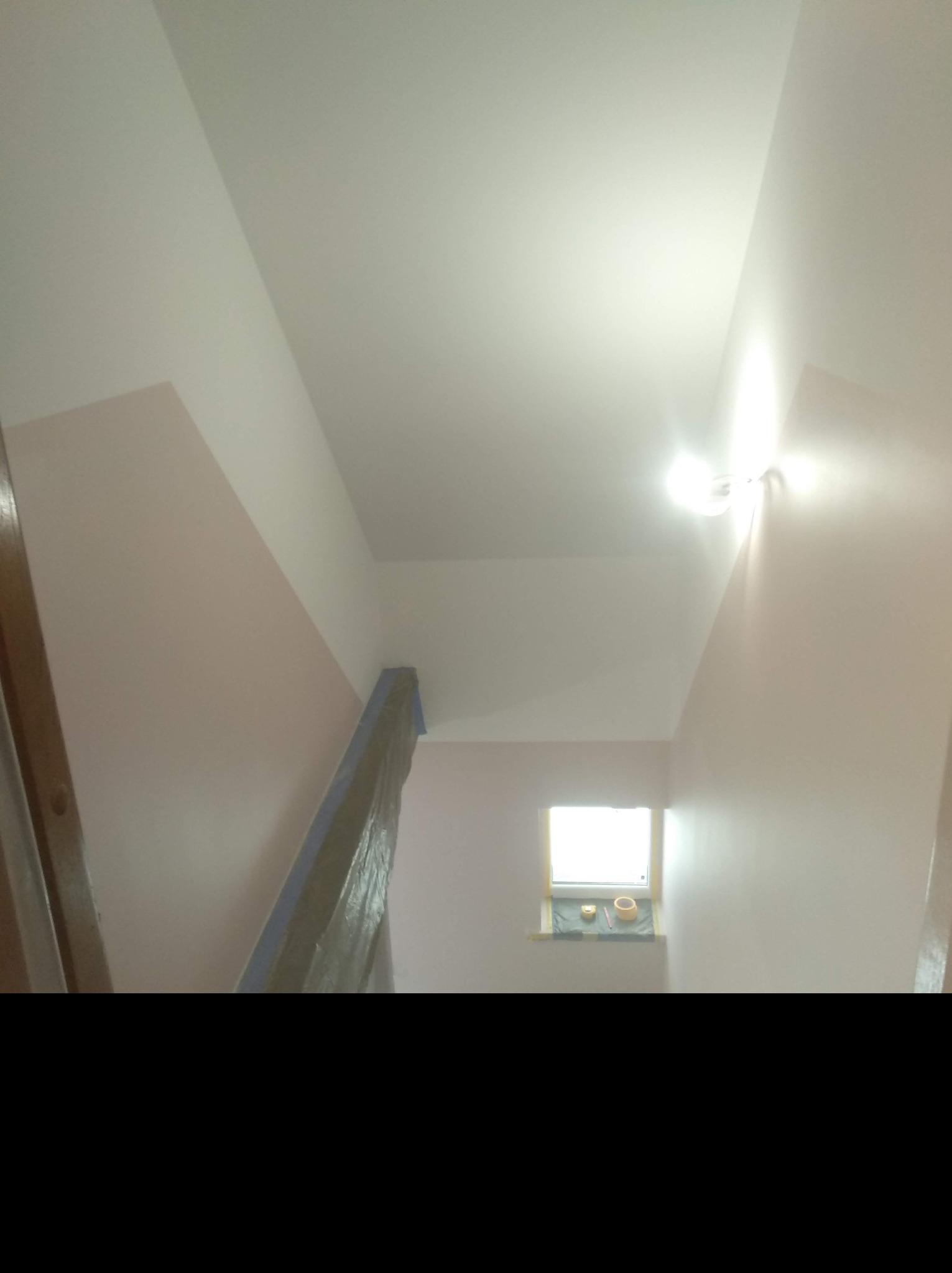 Klatka schodowa w trakcie malowania na jasny róż z białym sufitem, zabezpieczone schody, okno z akcesoriami malarskimi.