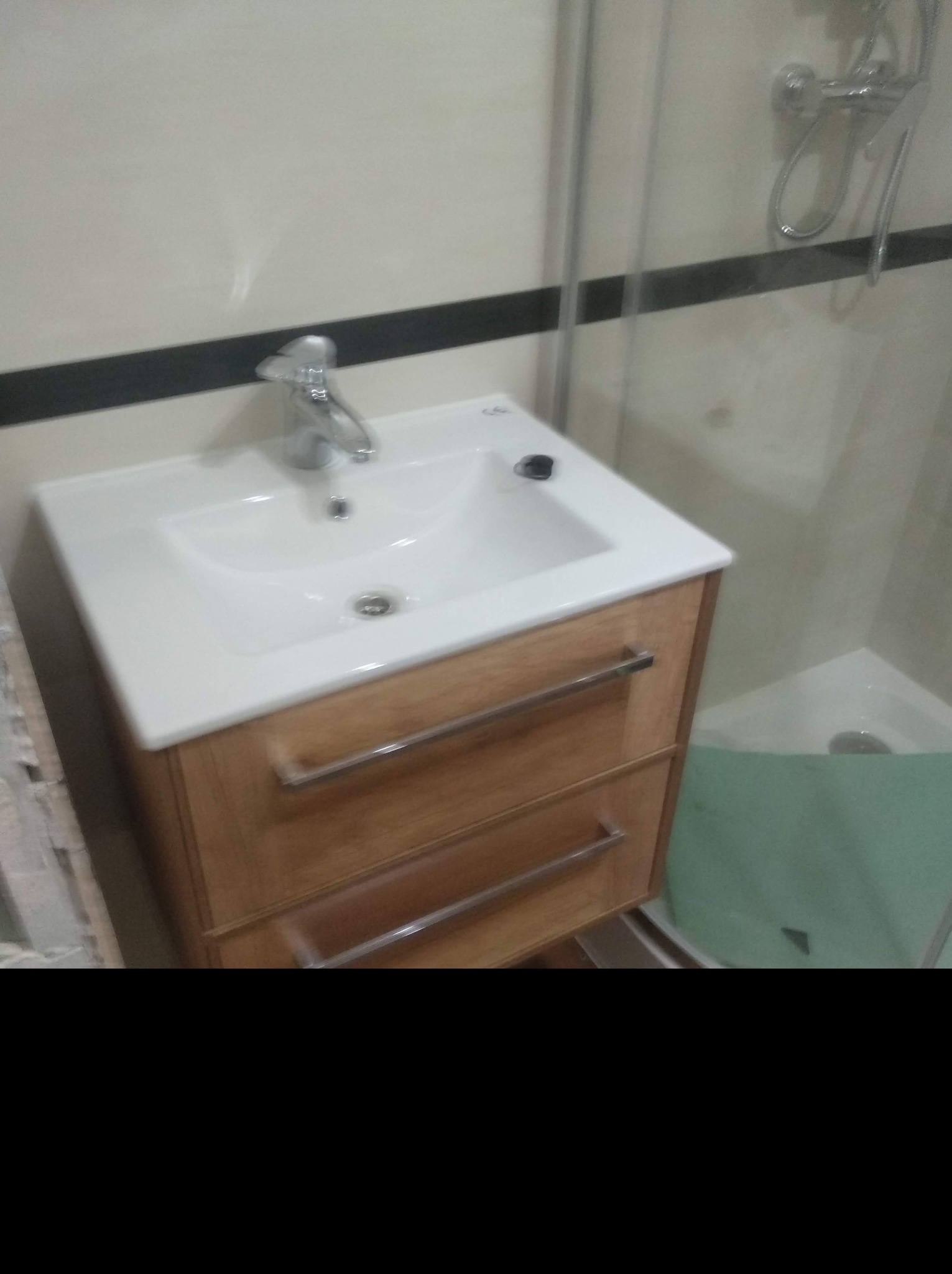 Biała umywalka z drewnianą szafką z dwoma szufladami w łazience z kabiną prysznicową w tle.