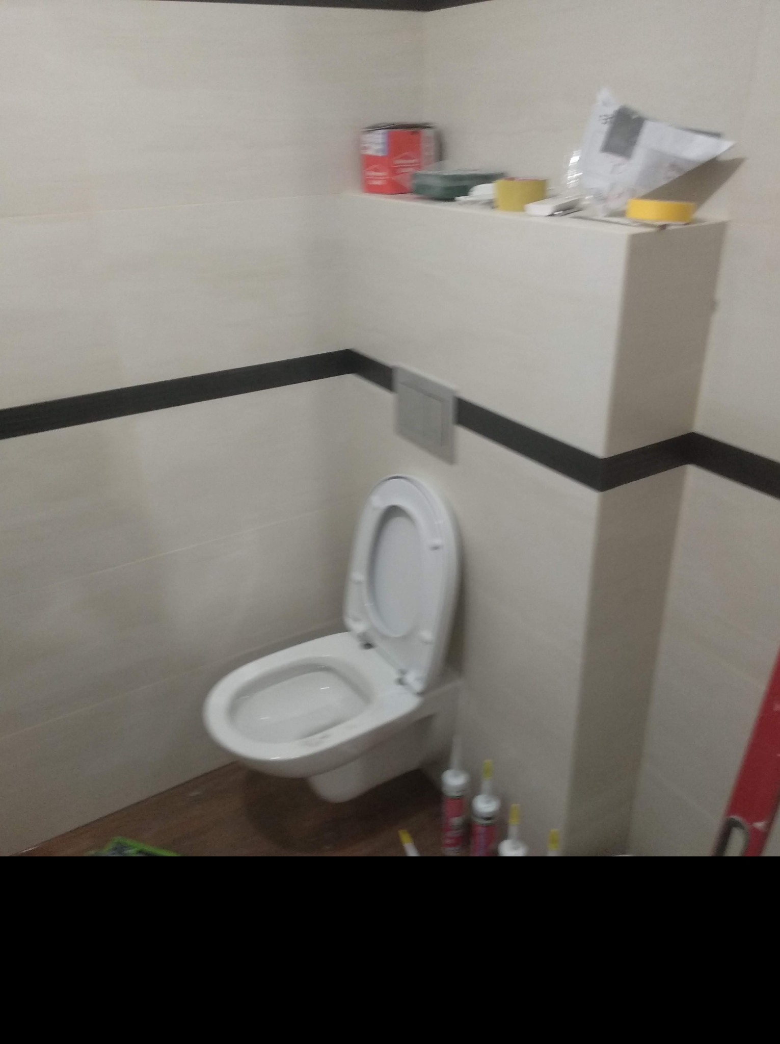 Zamontowana toaleta w łazience z białymi płytkami i czarnym pasem dekoracyjnym, widoczne silikony uszczelniające i narzędzia na półce.