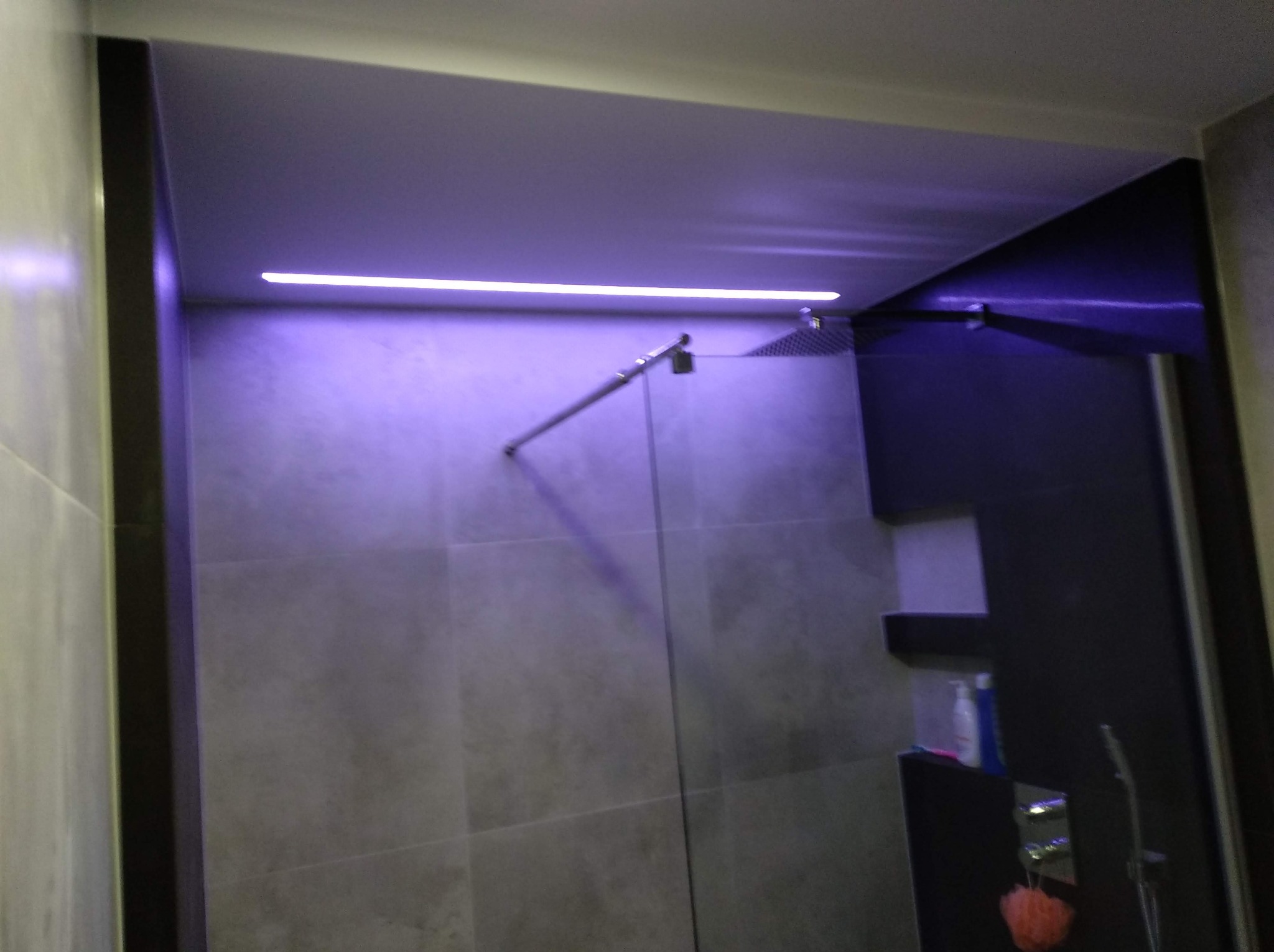 Nowoczesna kabina prysznicowa z deszczownicą, podświetlona listwą LED dającą fioletową poświatę, szare płytki na ścianach, czarne elementy wykończeniowe, widoczna półka z kosmetykami.