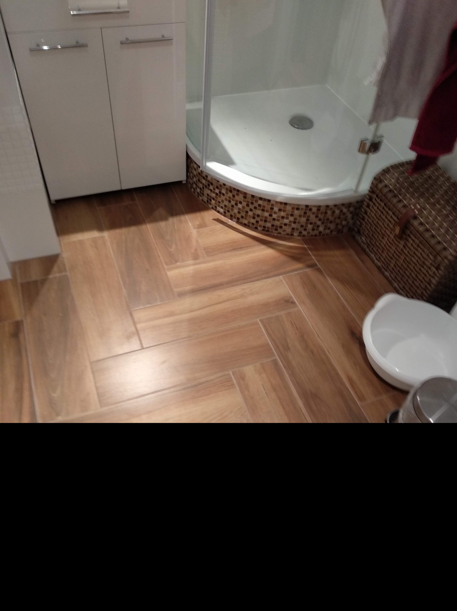 Wykończona łazienka z drewnopodobną podłogą ułożoną w jodełkę, kabiną prysznicową z mozaiką na podstawie i białą szafką pod umywalką. Widoczny kosz wiklinowy i biała miska.