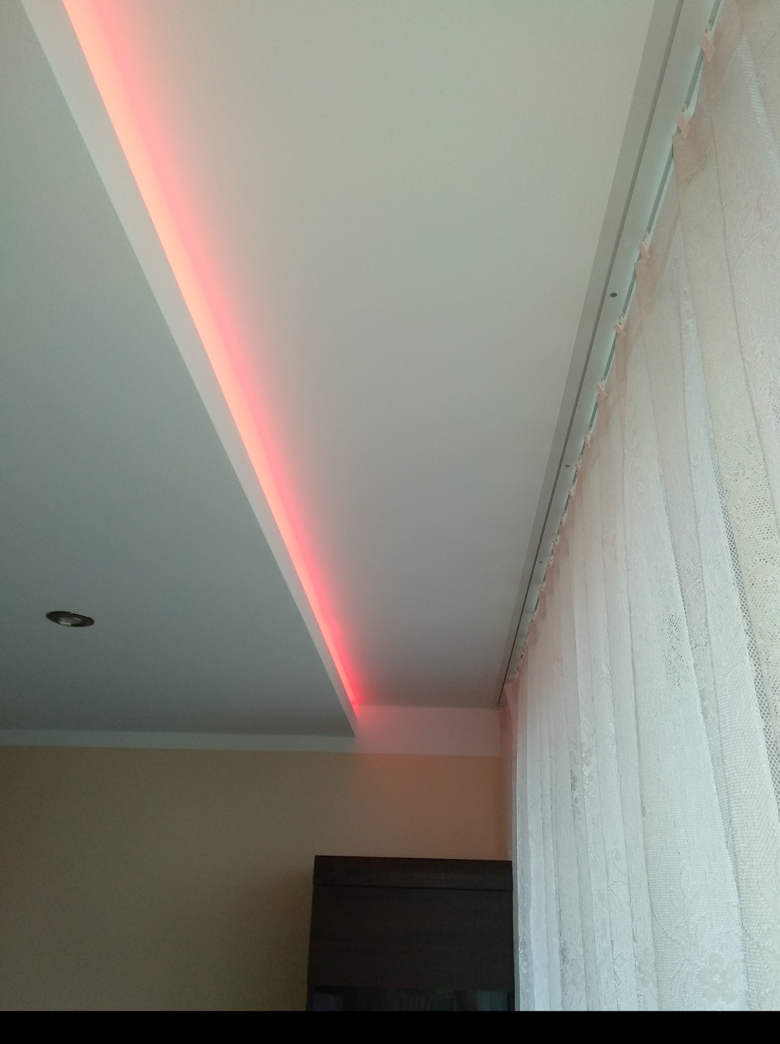 Podwieszany sufit z oświetleniem LED w kolorze czerwonym, widoczne firany i fragment mebla.