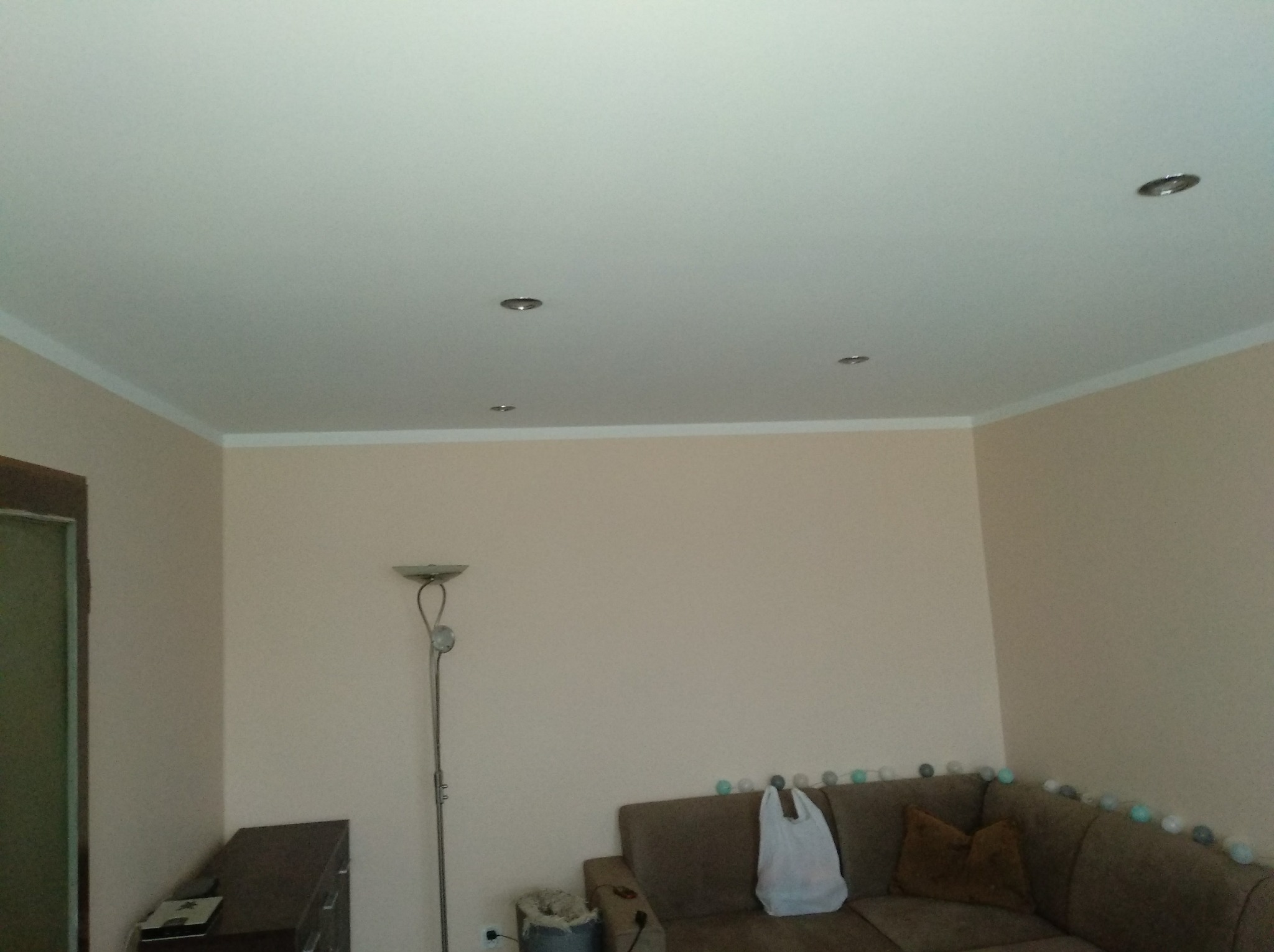 Jasny pokój po remoncie, widoczne gładkie ściany w odcieniach beżu, biały sufit z zamontowanymi halogenami i listwami przysufitowymi, częściowo widoczny narożnik sofy z dekoracyjnymi kulami...