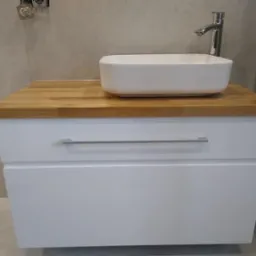 Biała szafka łazienkowa z dwiema szufladami, drewnianym blatem i nablatową umywalką w kształcie prostokąta, chromowana bateria, szare płytki na ścianie.