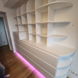 DrewnaMoc - Biała szafa wnękowa z półkami o zaokrąglonych krawędziach, podświetlona od dołu różowym światłem LED, zamontowana na drewnianej podłodze.