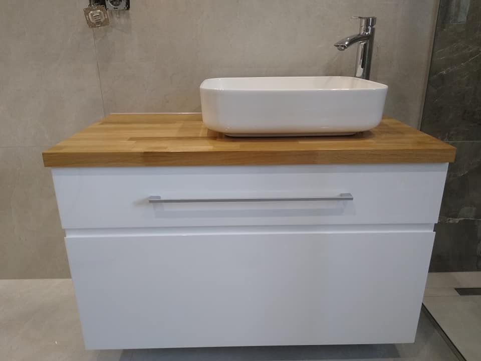 Biała szafka łazienkowa z dwiema szufladami, drewnianym blatem i nablatową umywalką w kształcie prostokąta, chromowana bateria, szare płytki na ścianie.