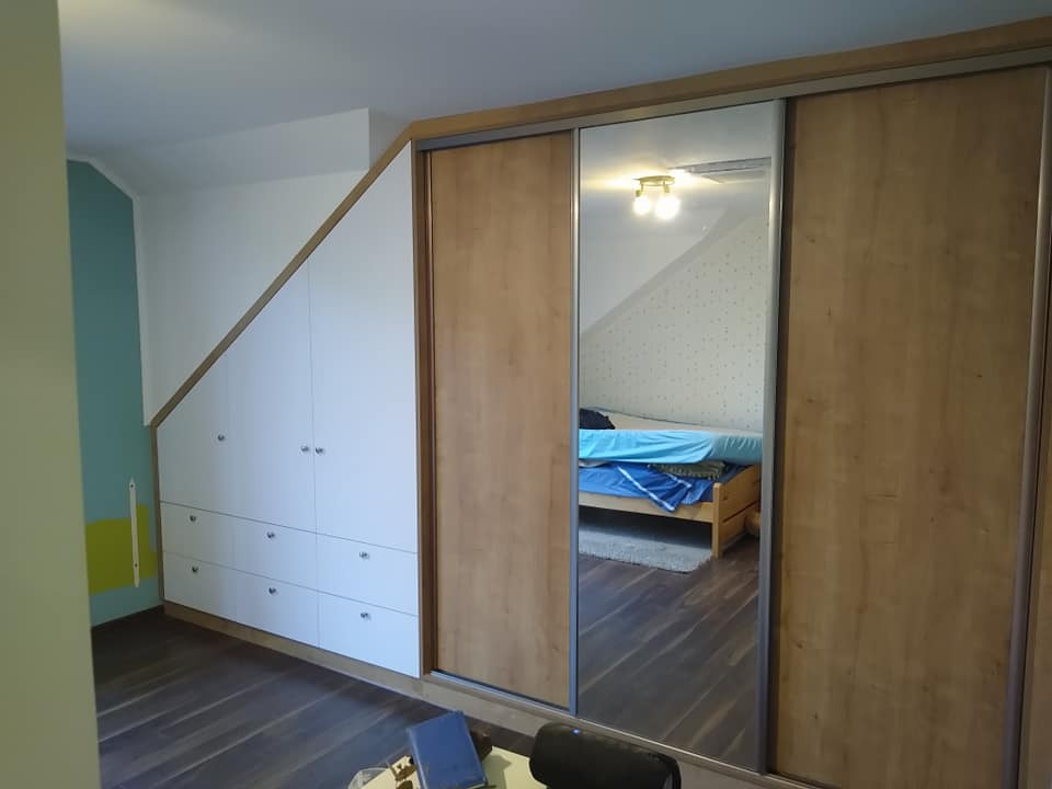 Zabudowa pod skosem ze szafkami i szufladami oraz szafa przesuwna z lustrem, odbijająca pokój z łóżkiem.