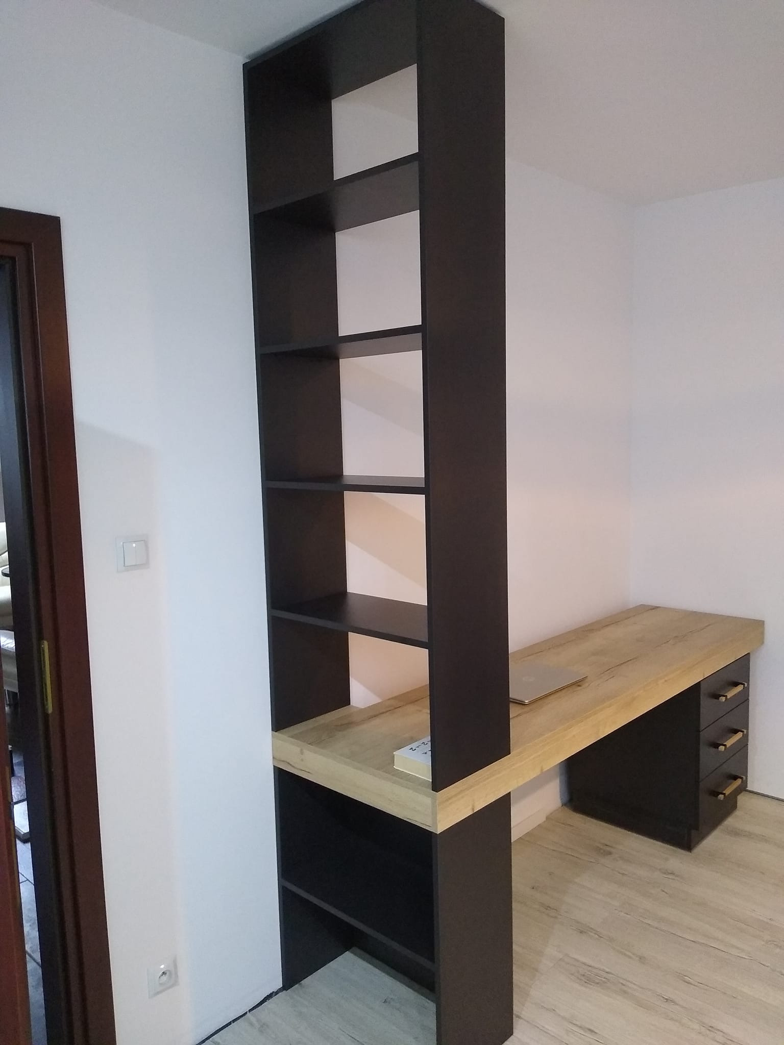 Ciemna, wysoka półka z biurkiem z blatem imitującym drewno, po lewej stronie drzwi, na biurku leży książka i laptop.