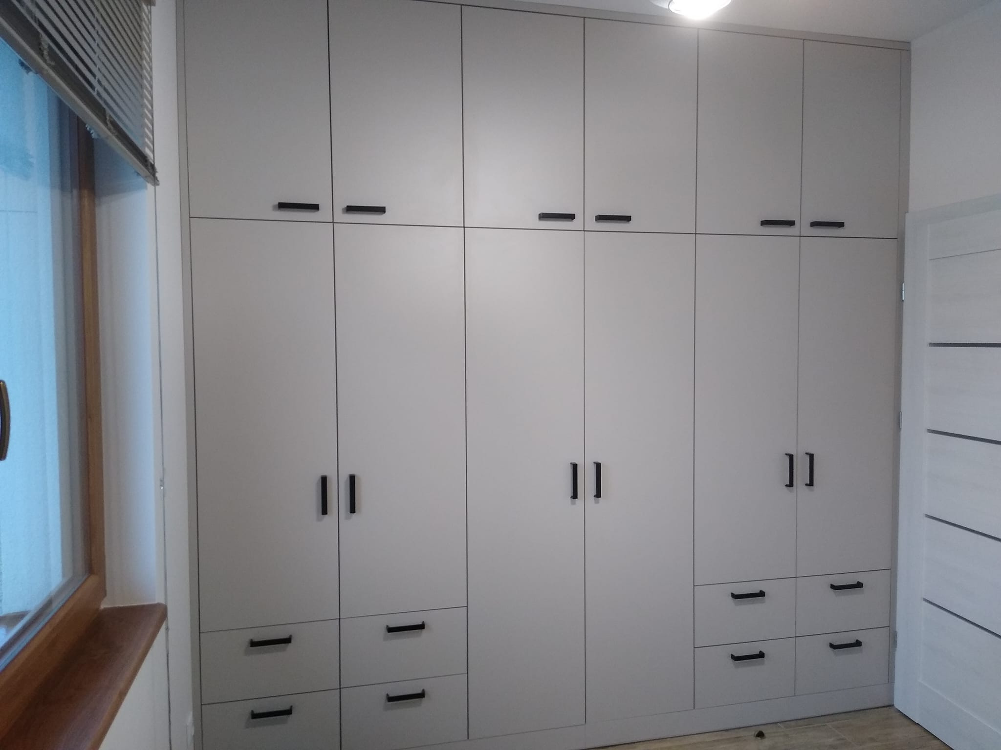 Duża, jasnoszara szafa wnękowa z czarnymi, podłużnymi uchwytami, zabudowana od podłogi do sufitu, widoczna obok okna z żaluzjami.