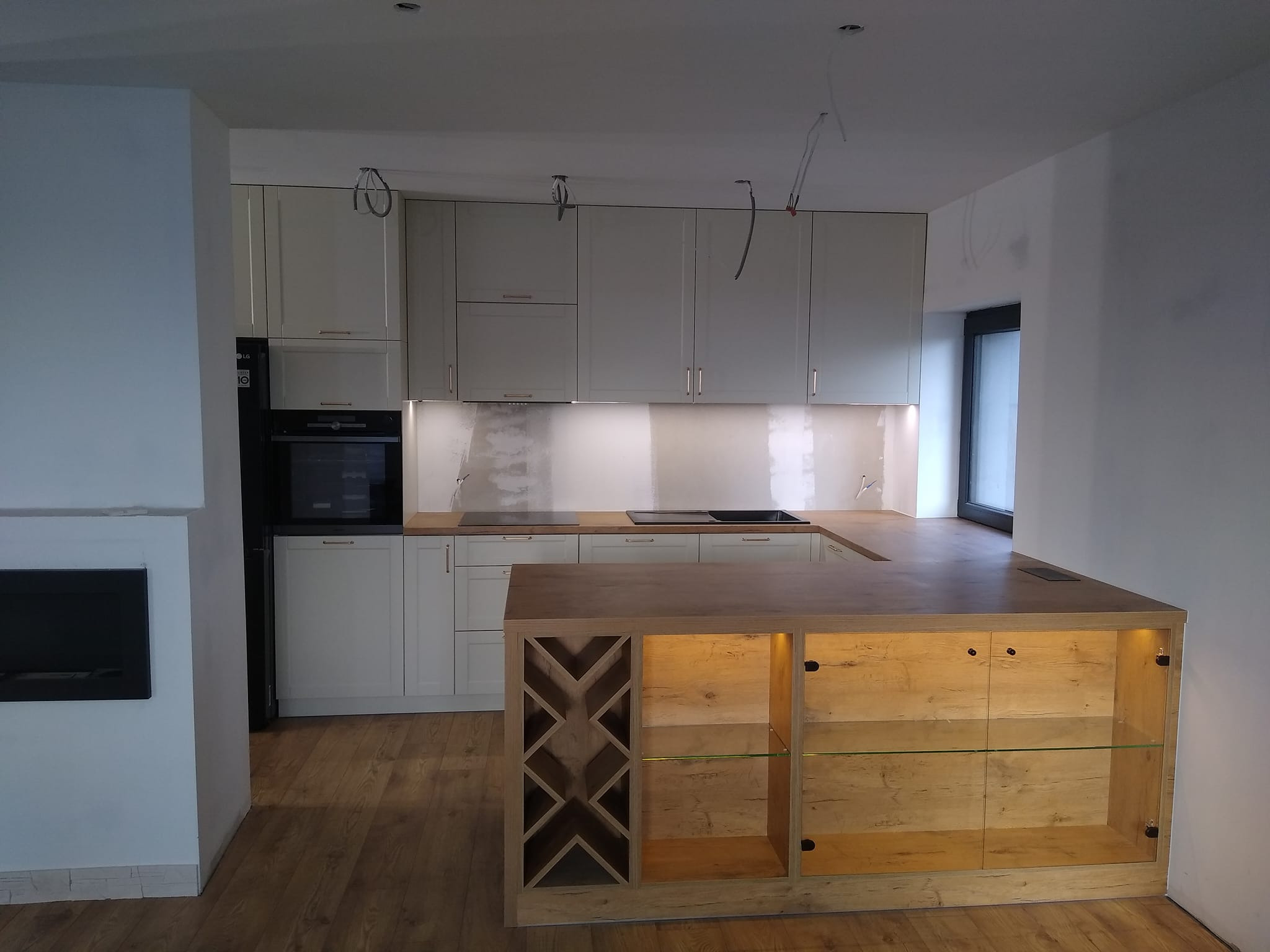 Kremowa zabudowa kuchenna z wyspą z wbudowanym oświetleniem i półkami na wino w Wilczkowicach, widoczne niedokończone prace wykończeniowe na ścianie i wiszące kable od oświetlenia.