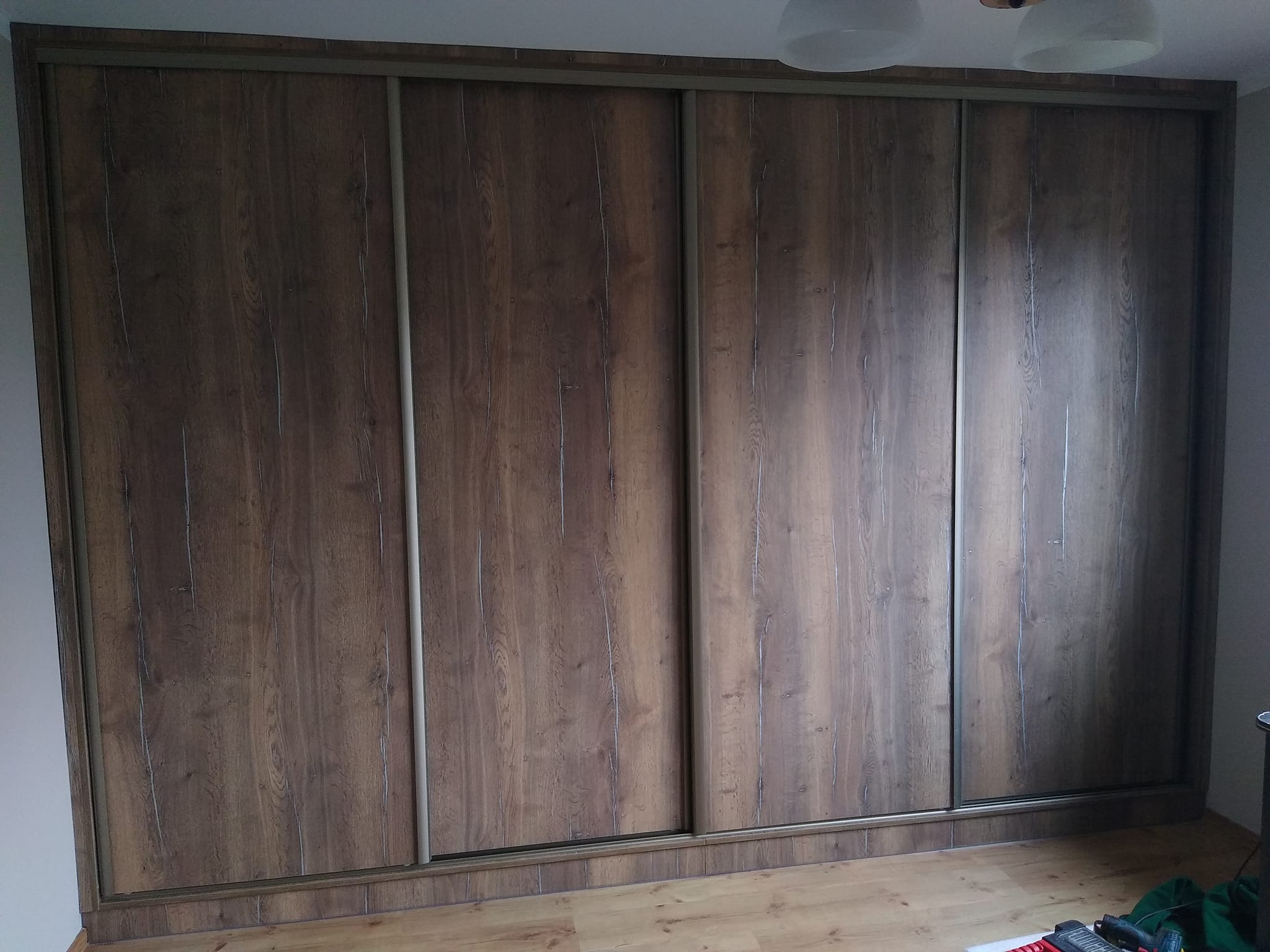 Duża szafa wnękowa z przesuwnymi drzwiami o wzorze ciemnego drewna i srebrnych prowadnicach, widoczna na tle parkietu.