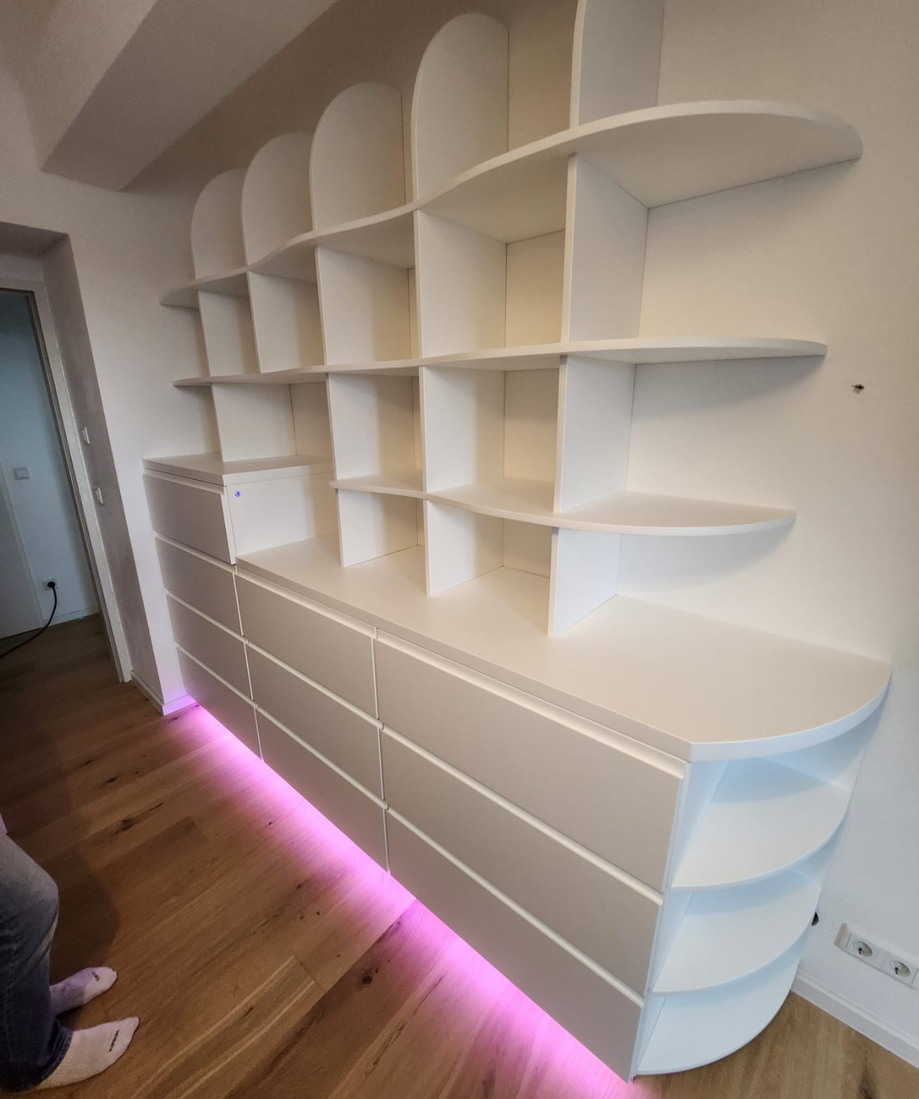 Biała szafa wnękowa z półkami o zaokrąglonych krawędziach, podświetlona od dołu różowym światłem LED, zamontowana na drewnianej podłodze.