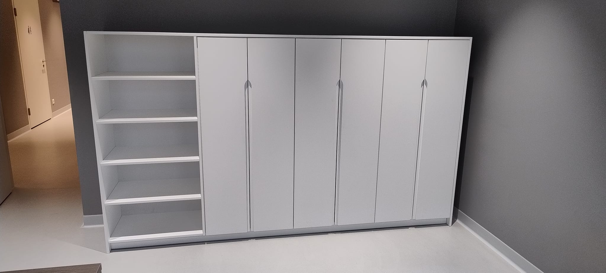 Biała szafa wnękowa z otwartymi półkami po lewej stronie, minimalistyczny design, widok z perspektywy, szara ściana w tle.