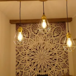 Trzy złote lampy wiszące w stylu industrialnym, z żarówkami Edisona, na tle ściany z dekoracyjnym, ażurowym wzorem mandali i drewnianej belki stropowej.