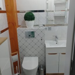 Danio - Nowoczesna łazienka z wiszącą toaletą, umywalką z szafką i grzejnikiem drabinkowym, wykończona białymi płytkami z subtelnym wzorem i drewnianym elementem konstrukcyjnym.