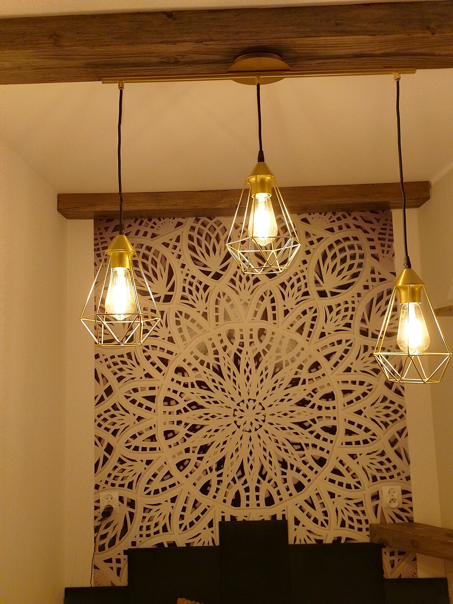 Trzy złote lampy wiszące w stylu industrialnym, z żarówkami Edisona, na tle ściany z dekoracyjnym, ażurowym wzorem mandali i drewnianej belki stropowej.