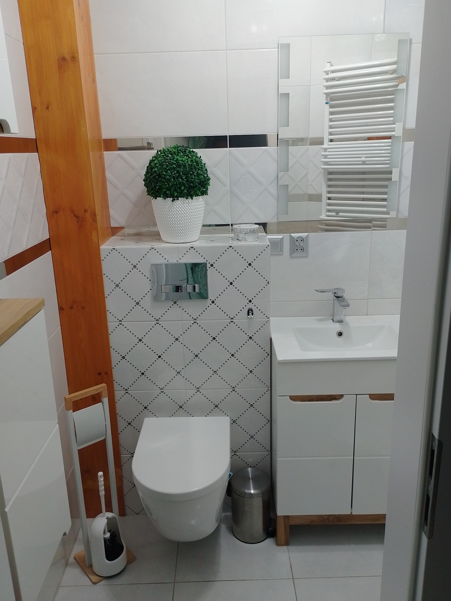 Nowoczesna łazienka z wiszącą toaletą, umywalką z szafką i grzejnikiem drabinkowym, wykończona białymi płytkami z subtelnym wzorem i drewnianym elementem konstrukcyjnym.