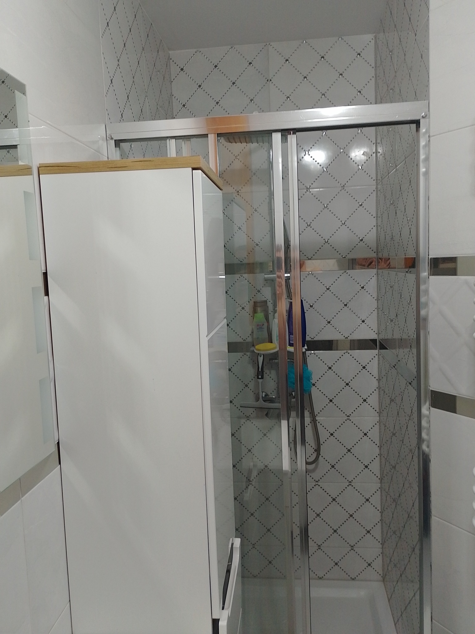 Biała szafka łazienkowa obok kabiny prysznicowej z przesuwnymi drzwiami i biało-szarą mozaiką na ścianach.