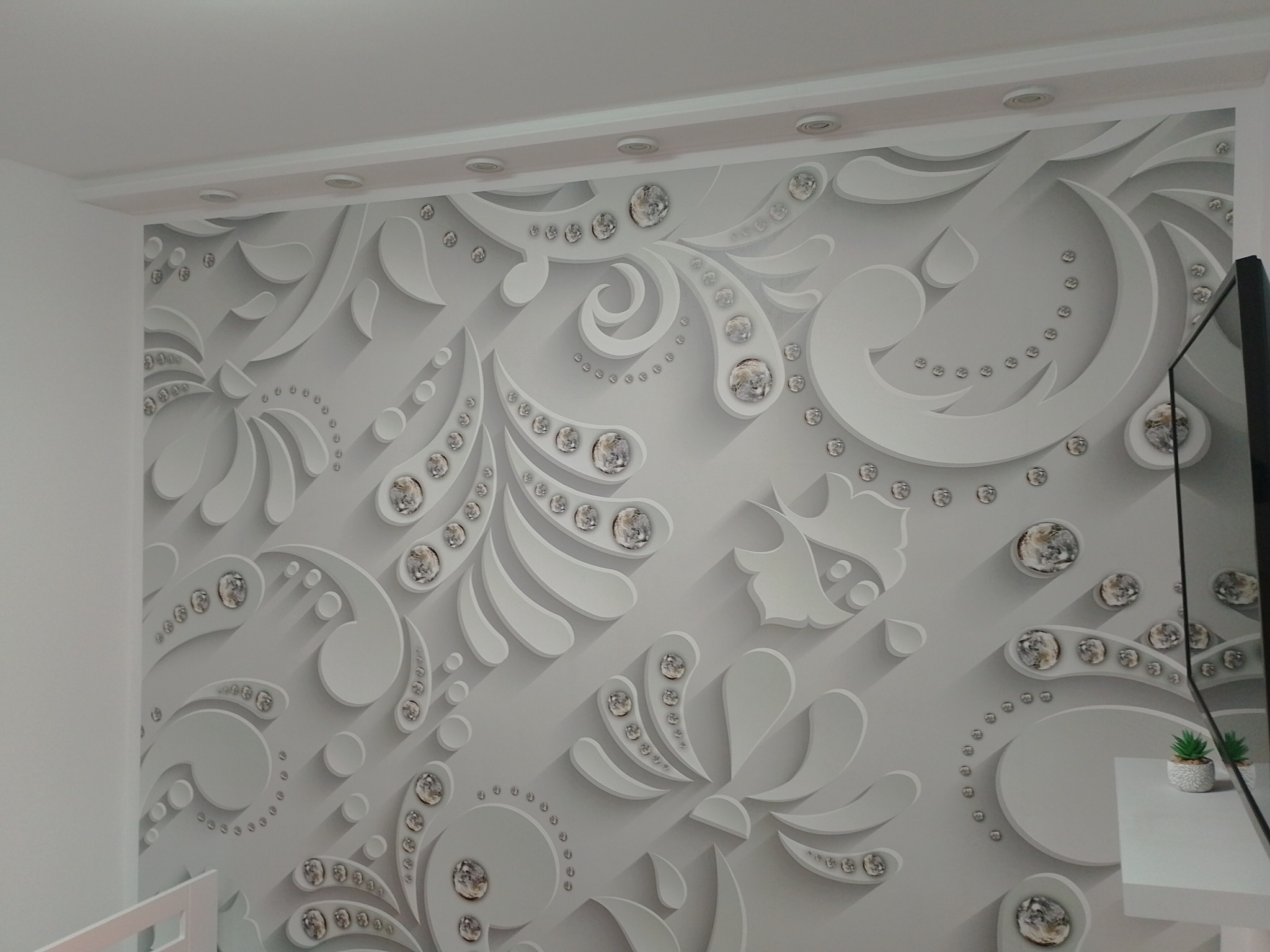 Biała ściana z trójwymiarowym, dekoracyjnym wzorem w stylu ornamentów, ozdobiona elementami przypominającymi kryształki, oświetlona przez wbudowane lampy sufitowe.