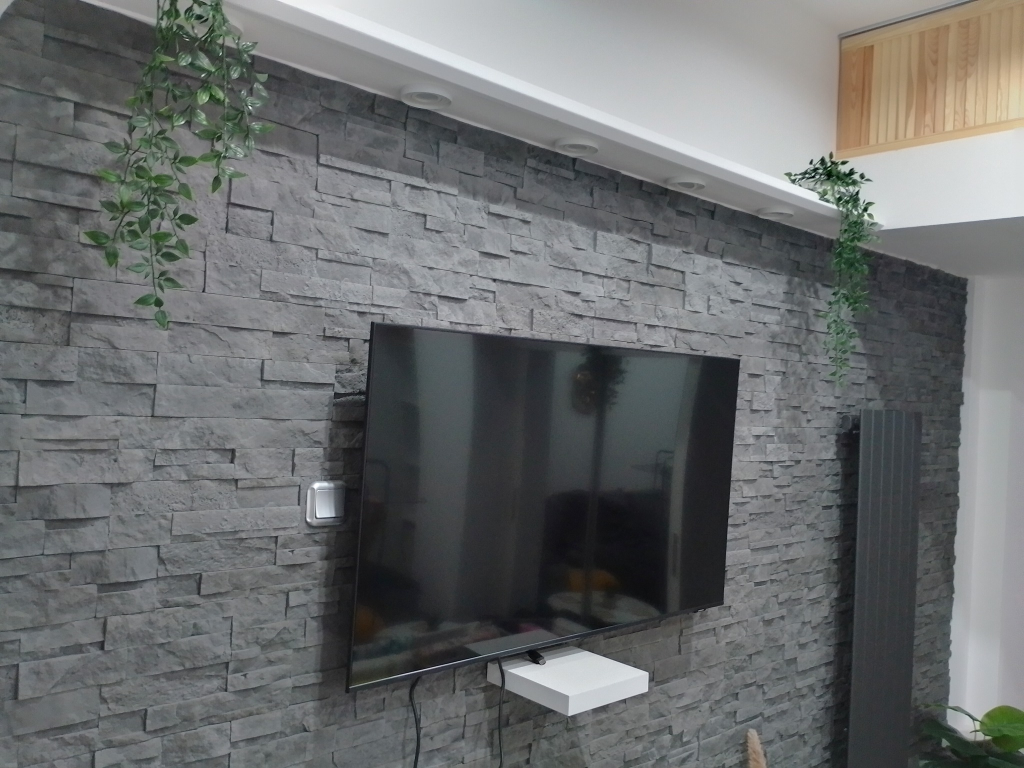 Telewizor zawieszony na ścianie wyłożonej szarym kamieniem dekoracyjnym z wiszącymi sztucznymi roślinami po bokach, biała półka pod telewizorem.