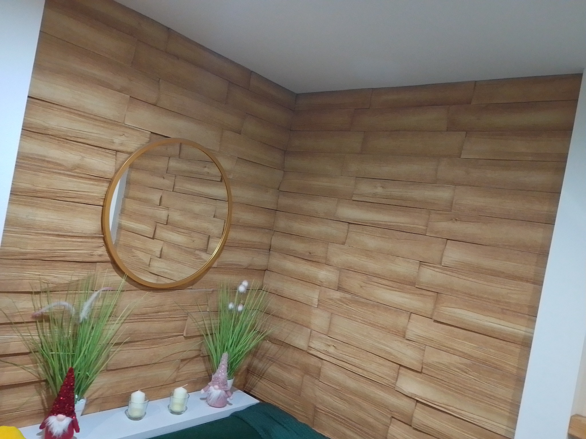 Ściana wyłożona panelami imitującymi drewno, okrągłe lustro w złotej ramie, dekoracje roślinne i figurki na półce
