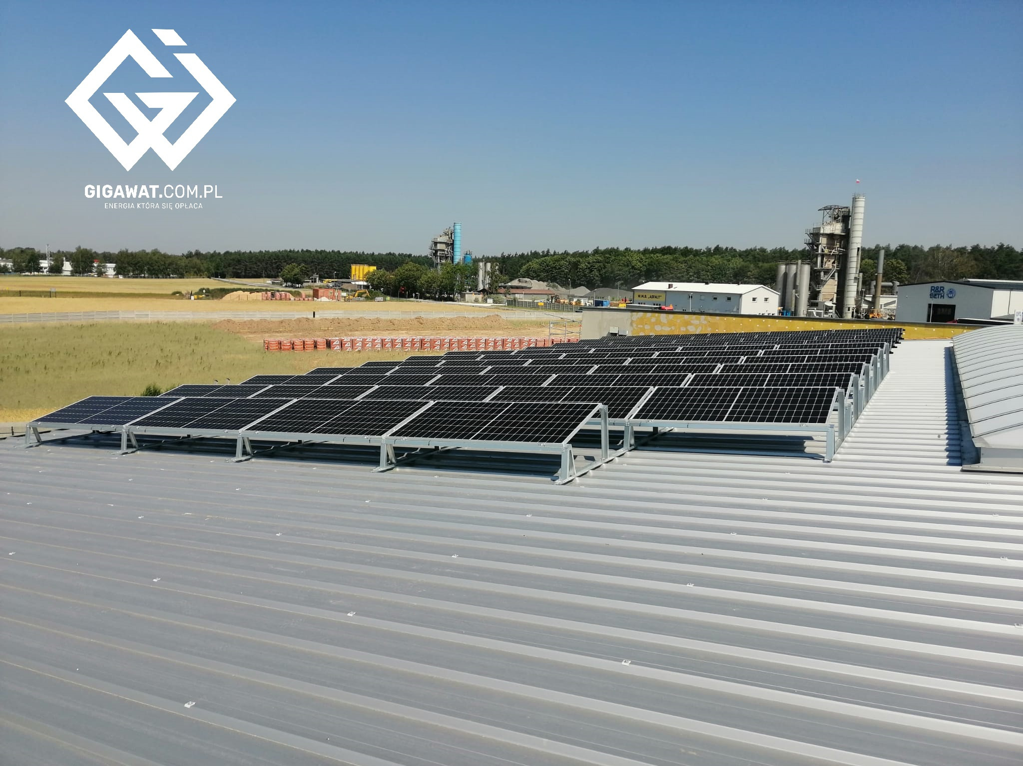 Rząd paneli słonecznych na metalowym dachu przemysłowym, z logo firmy GIGAWAT.COM.PL w górnym lewym rogu i widokiem na fabrykę w tle.