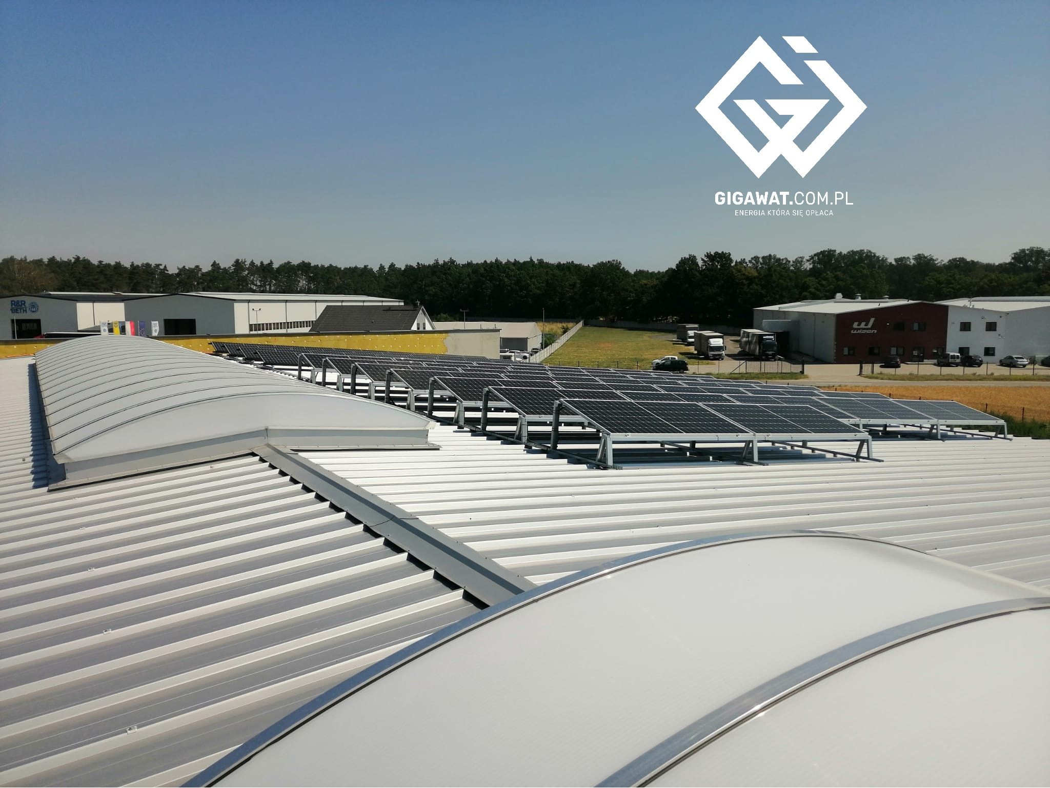 Rząd paneli słonecznych zamontowanych na dachu przemysłowym z świetlikami dachowymi, widok z góry, w tle budynki przemysłowe i drzewa.