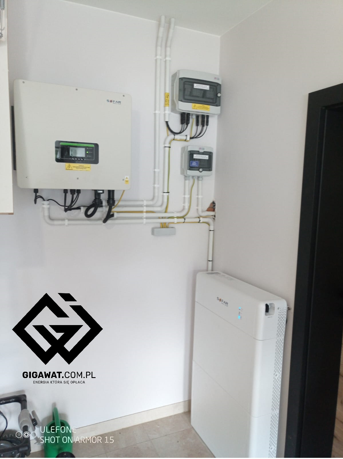 Zainstalowany falownik hybrydowy Sofar oraz magazyn energii Sofar BTS 10kWh z modułem zasilania rezerwowego, widoczne białe rury instalacyjne i skrzynki elektryczne na białej ścianie w pomieszczeniu.