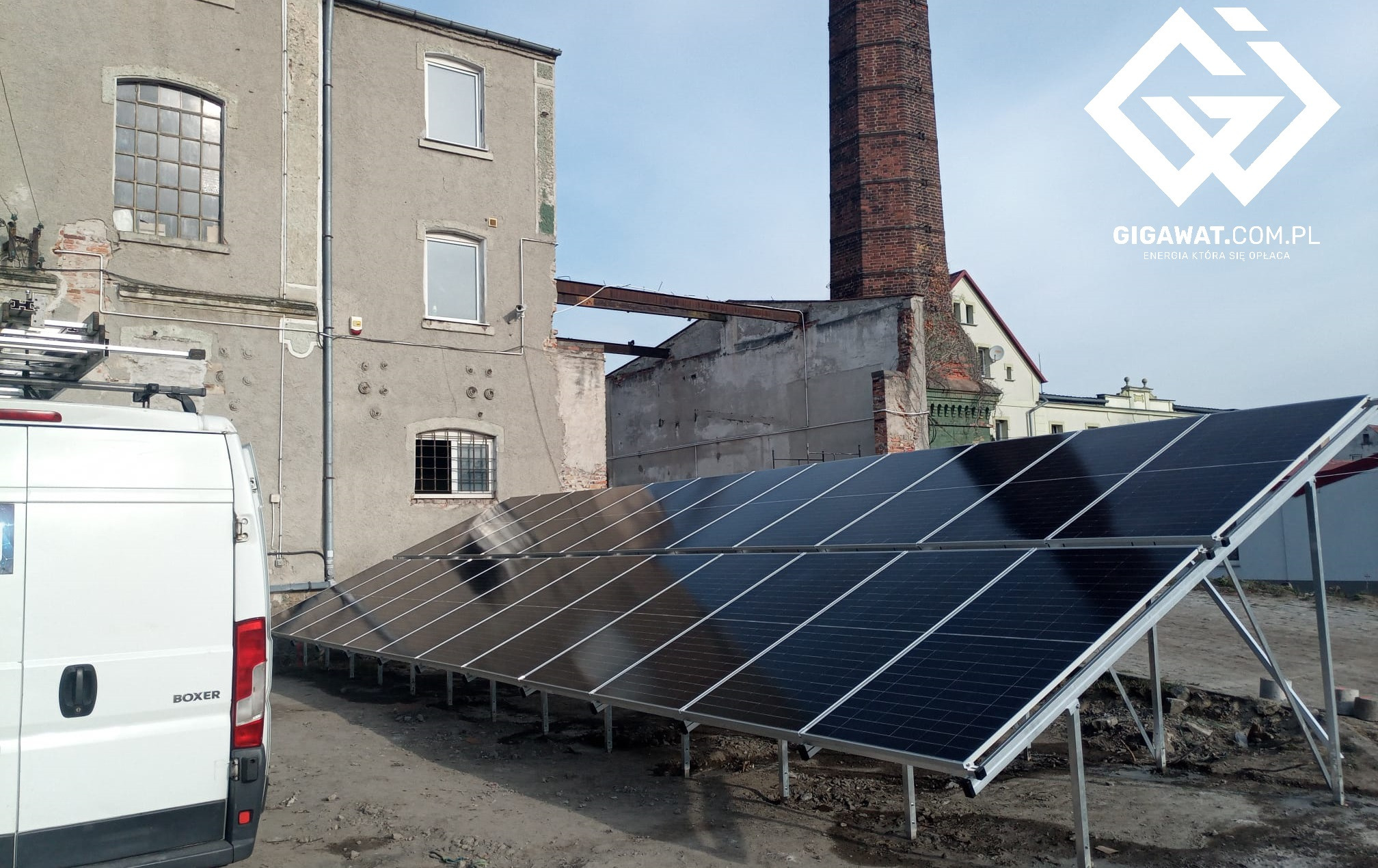 Instalacja paneli słonecznych na dachu budynku przemysłowego z ceglanym kominem w tle, obok zaparkowany biały furgon.