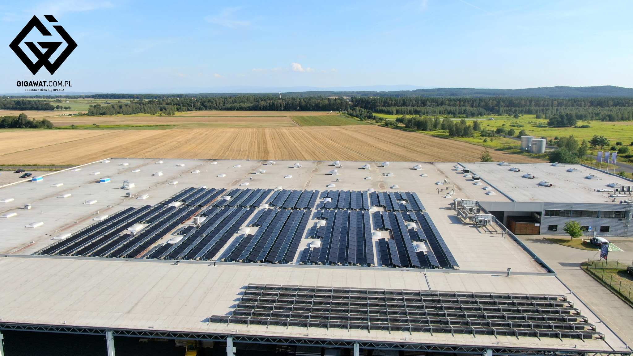 Widok z góry na dużą instalację paneli słonecznych na dachu hali produkcyjnej, z logo firmy GIGAWAT.COM.PL w lewym górnym rogu, otoczoną polami i lasami w województwie dolnośląskim.