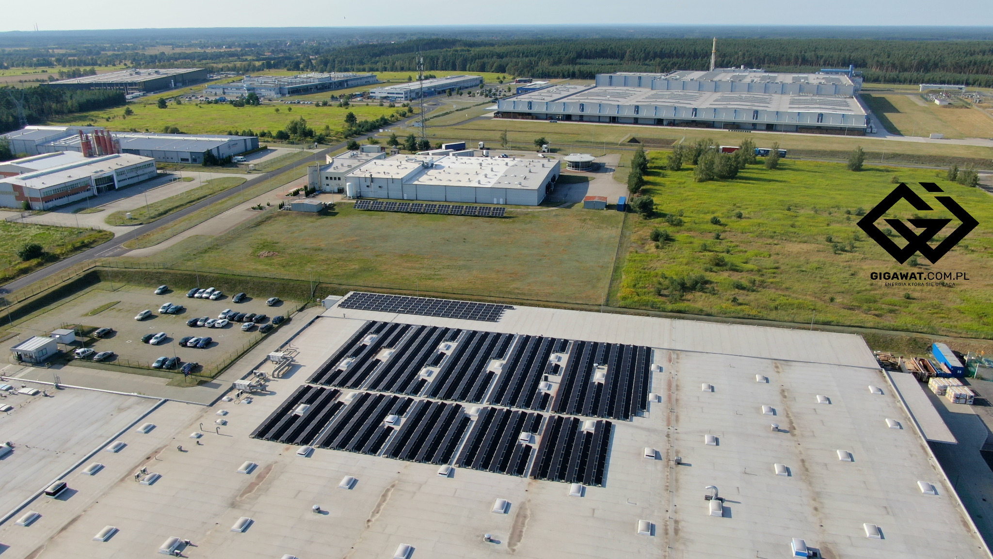 Widok z lotu ptaka na rozległą instalację paneli słonecznych na dachu hali produkcyjnej, z widocznym logo firmy na tle krajobrazu przemysłowego i leśnego w oddali.