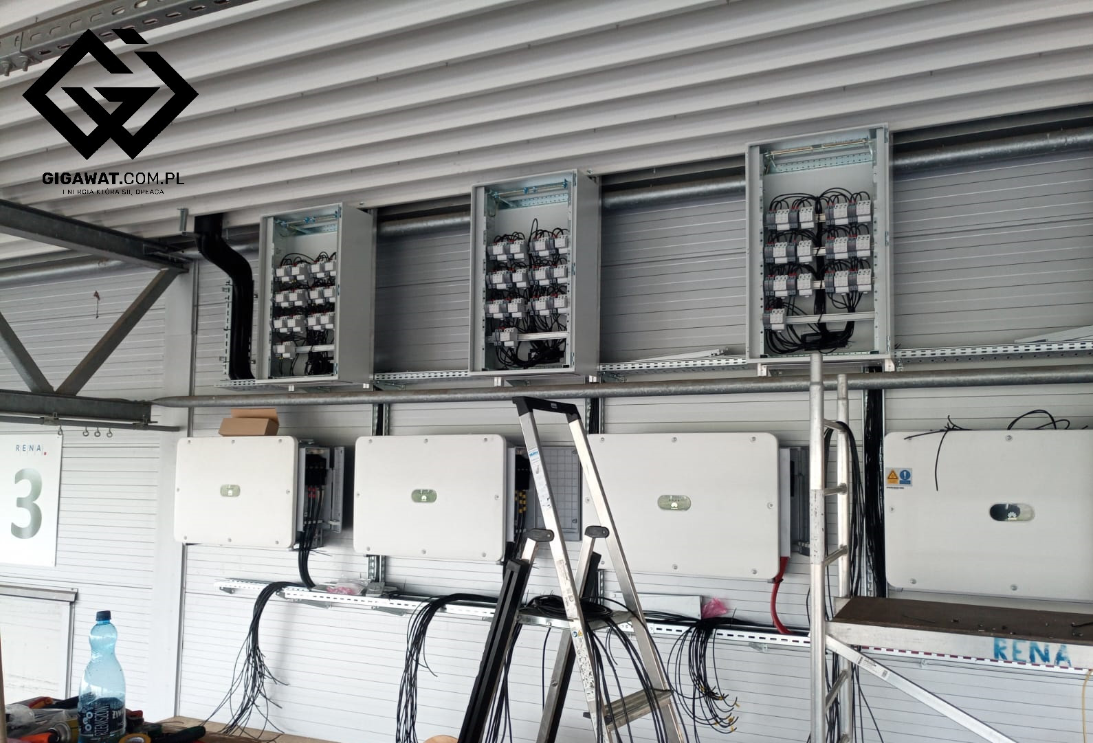 Wnętrze hali produkcyjnej z zamontowanymi czterema białymi inwerterami Huawei 100kW na ścianie, powyżej widoczne otwarte skrzynki rozdzielcze z okablowaniem, częściowo zasłonięte przez rozłożoną...