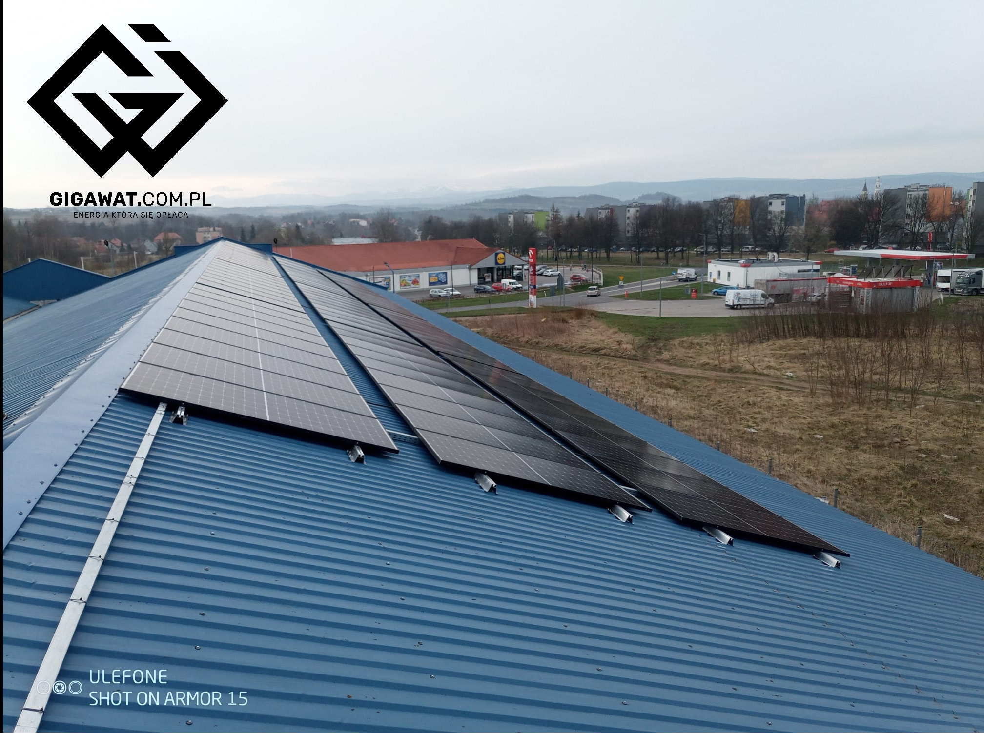 Instalacja paneli na blaszanym dachu budynku przemysłowego, widok z góry na panele i otoczenie miejskie z budynkami i drzewami w tle, pochmurne niebo.