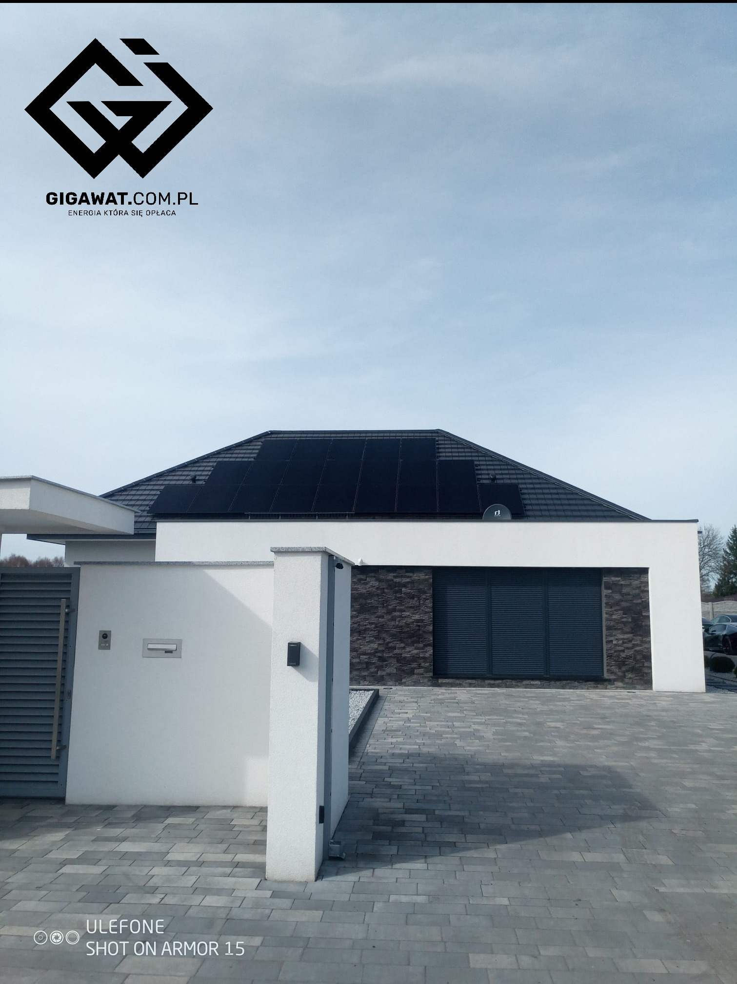 Czarny dach nowoczesnego domu jednorodzinnego z panelami fotowoltaicznymi full black w technologii podwójnego szkła, widok z poziomu podjazdu z kostki brukowej, logo firmy Gigawat.com.pl w lewym...