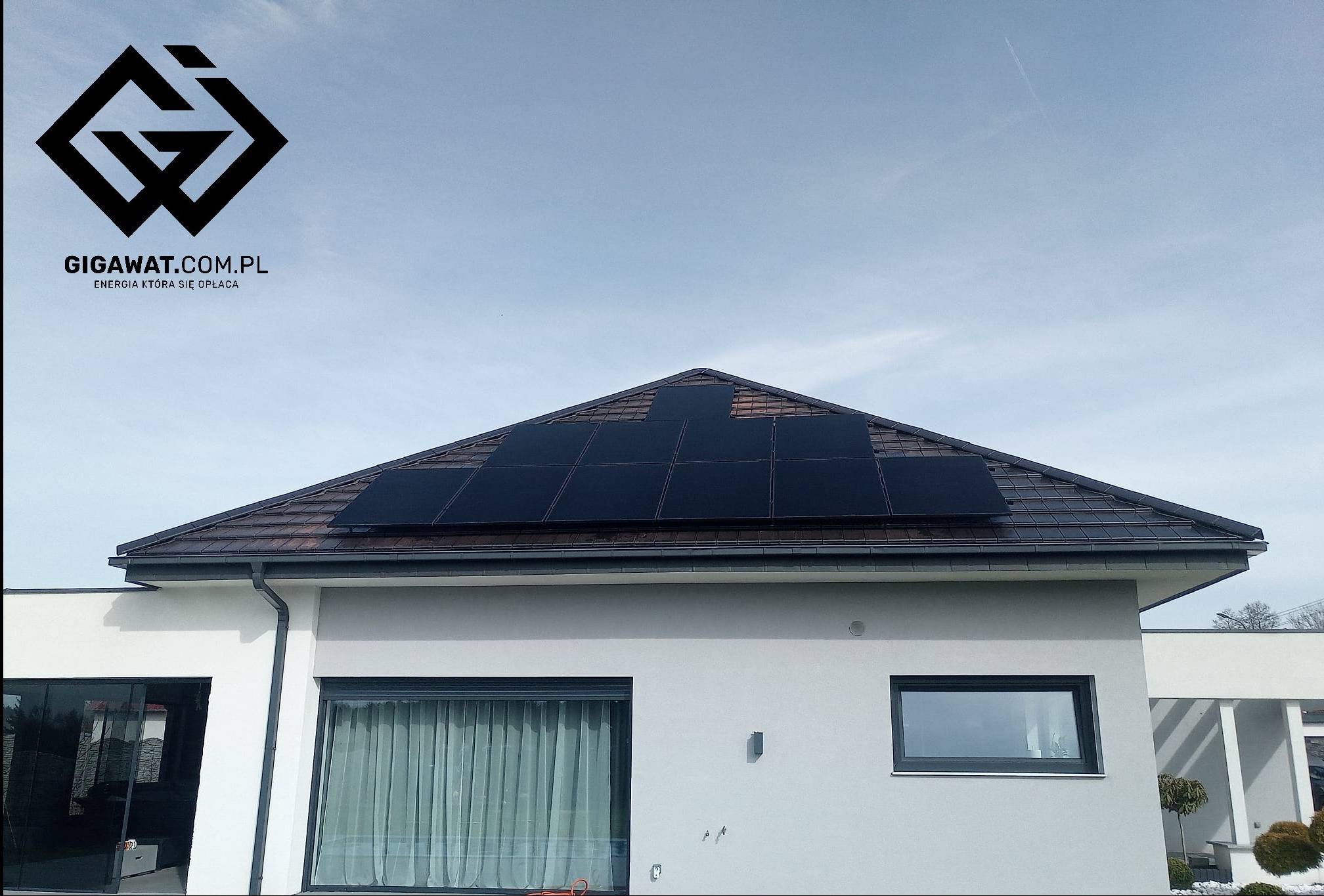 Czarny panel na brązowym dachu domu jednorodzinnego z logo firmy GIGAWAT.COM.PL na niebie.