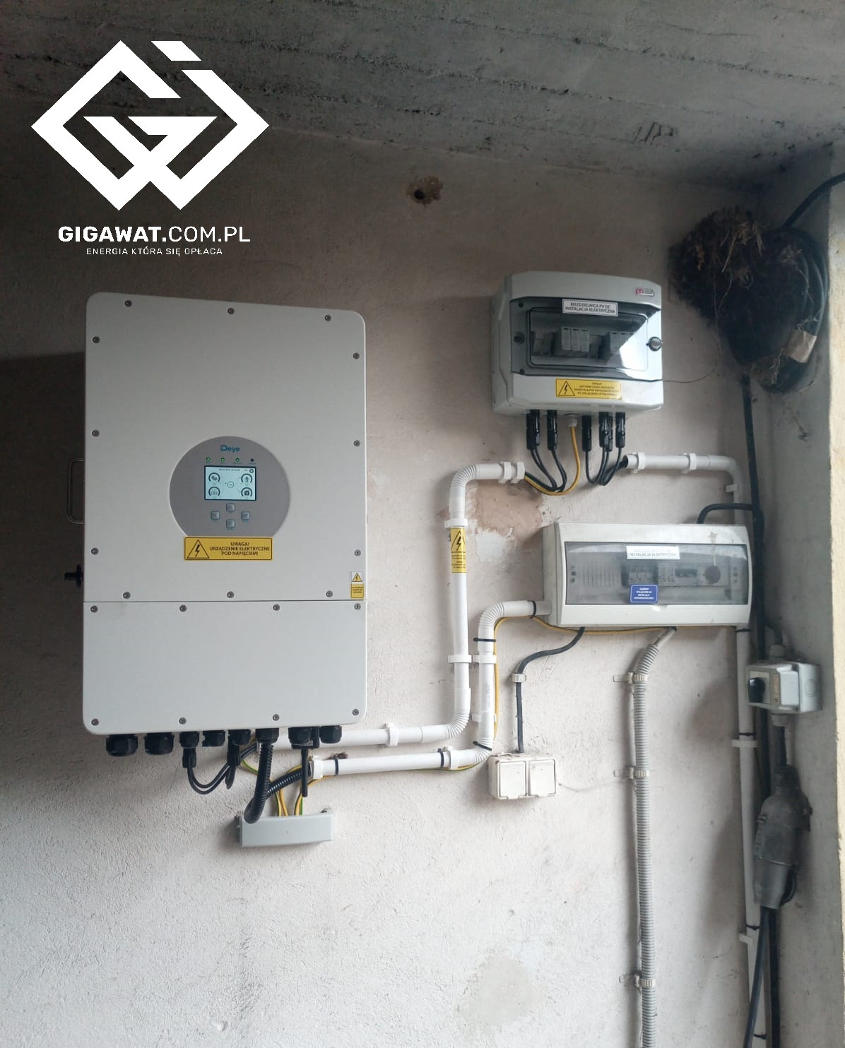Szary inwerter z podłączonymi kablami, skrzynki elektryczne i rurki PCV na nierównej, białej ścianie wewnątrz budynku. U góry logo firmy.