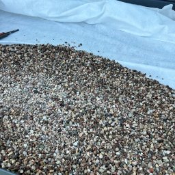 DACHY, TARASY, BALKONY, PIOTR BURLITA - Warstwa drobnego, kolorowego żwiru na białej geowłókninie, częściowo przykryta ręką w roboczej rękawicy, widoczny fragment okna i nóż do cięcia, przygotowanie do prac dekarskich.