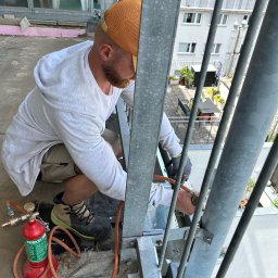 DACHY, TARASY, BALKONY, PIOTR BURLITA - Dekarz w trakcie pracy na balkonie, używający palnika do obróbki blacharskiej, widoczne narzędzia i elementy konstrukcji balkonu.