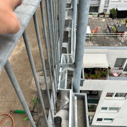 DACHY, TARASY, BALKONY, PIOTR BURLITA - Widok z góry na fragment dachu z zamontowanymi metalowymi rynnami, widoczna ręka na balustradzie, narzędzia dekarskie i fragment elewacji budynku z balkonami.