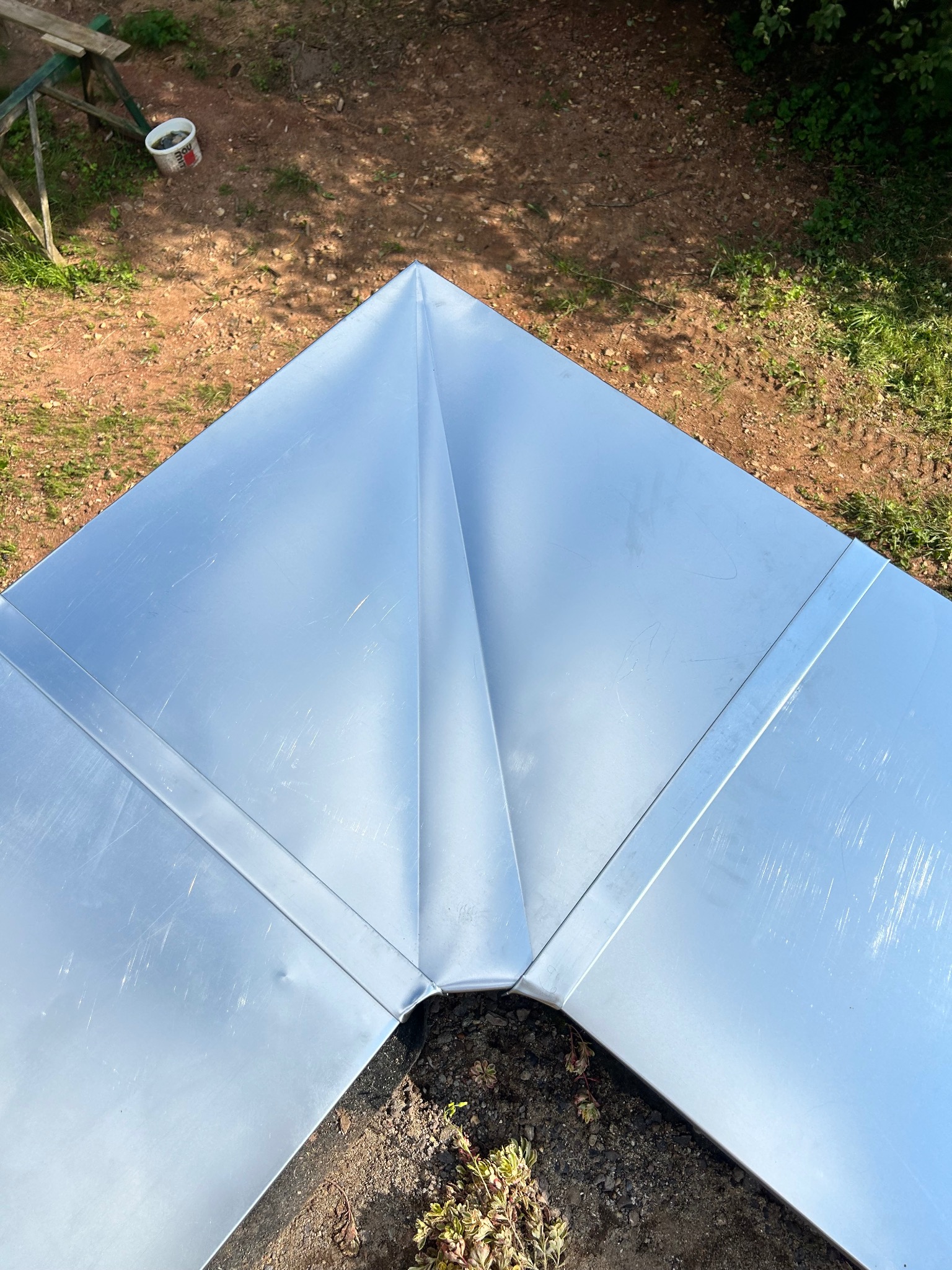 Metalowy element dachu z wyraźnym łączeniem blach na szczycie, widoczne zarysowania na powierzchni, w tle ziemia i roślinność.