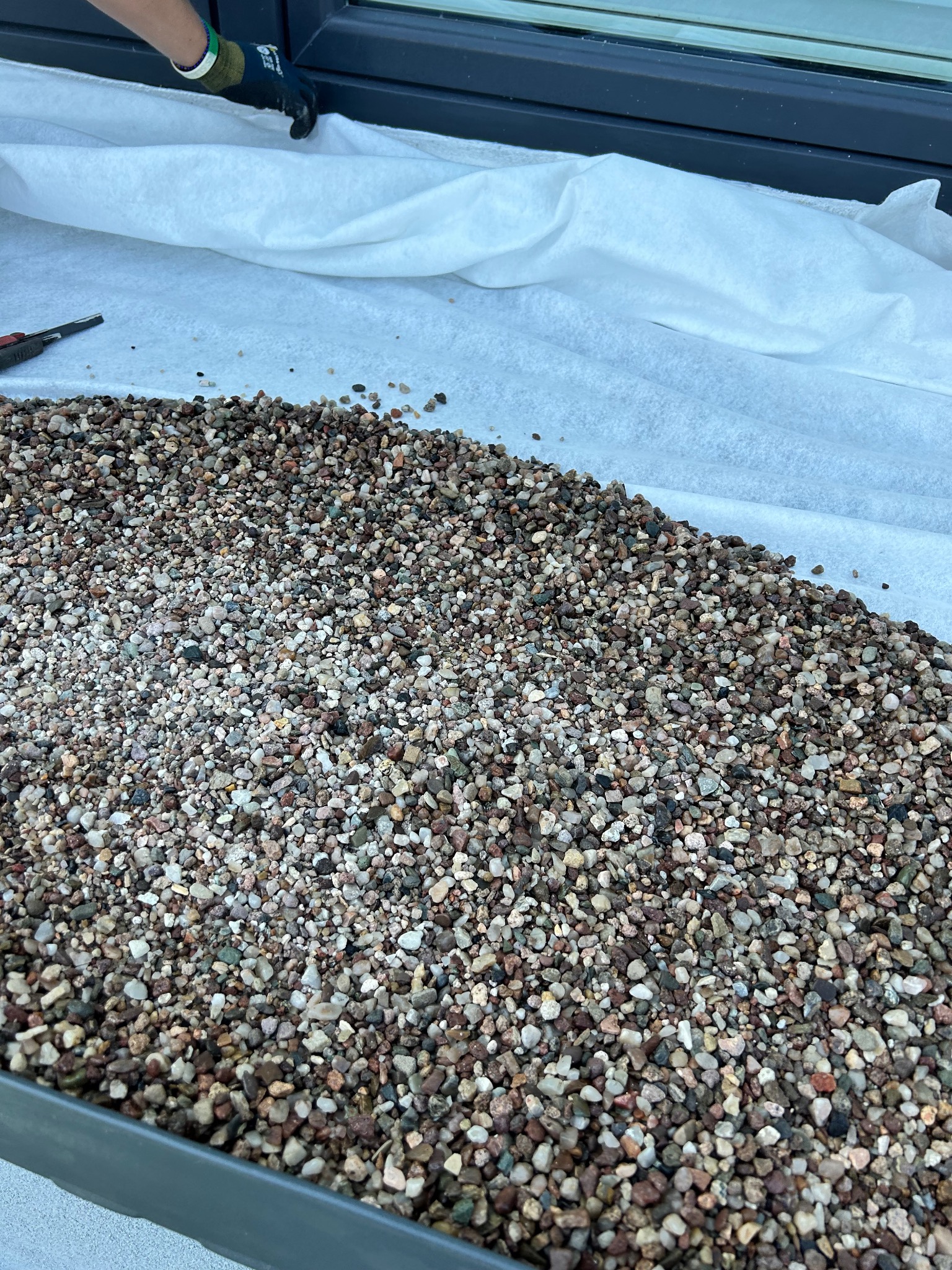 Warstwa drobnego, kolorowego żwiru na białej geowłókninie, częściowo przykryta ręką w roboczej rękawicy, widoczny fragment okna i nóż do cięcia, przygotowanie do prac dekarskich.