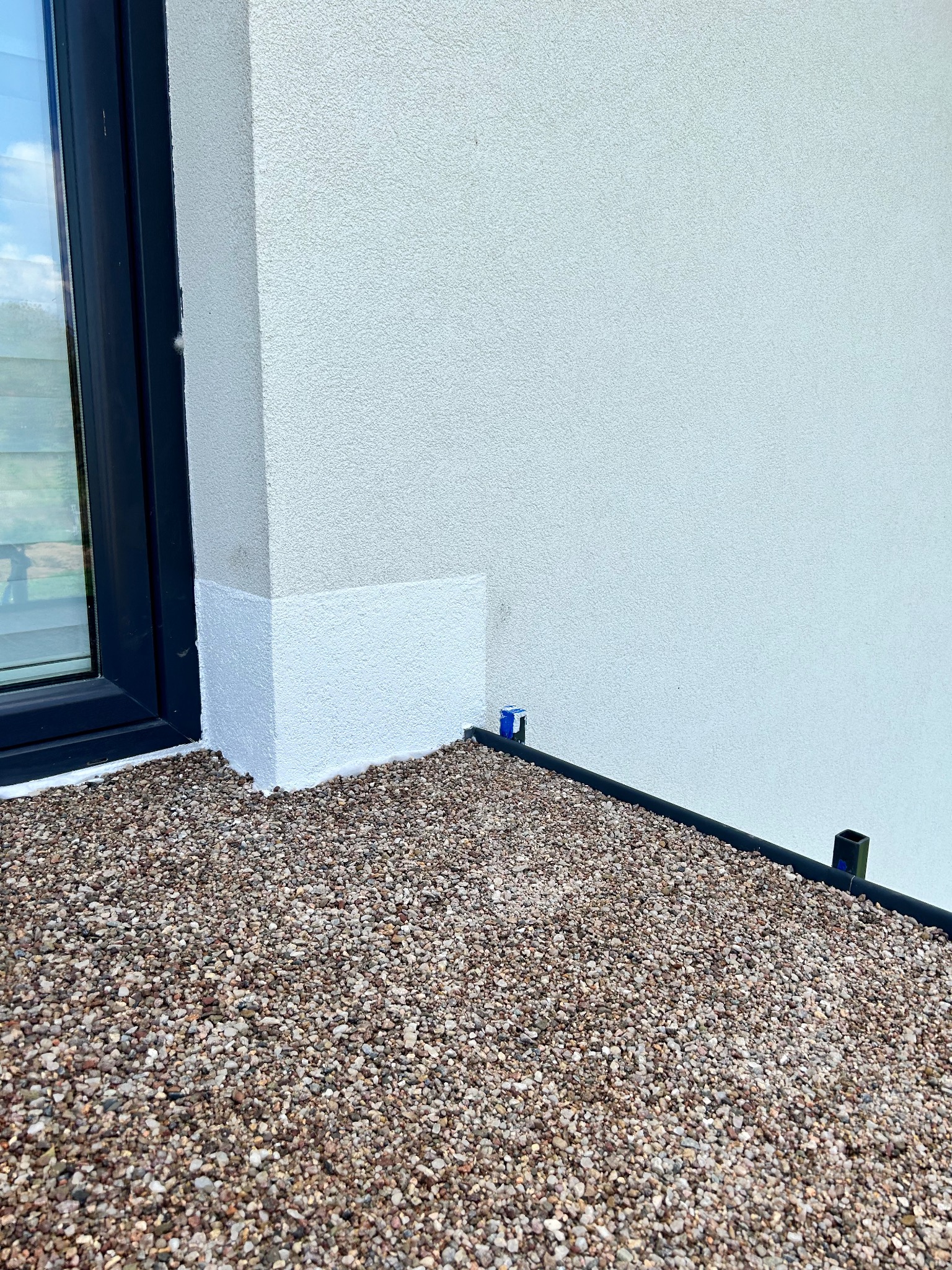 Szczegółowe ujęcie ściany budynku z wykończeniem z drobnych kamyczków i czarną listwą przypodłogową oraz fragment okna po lewej stronie.