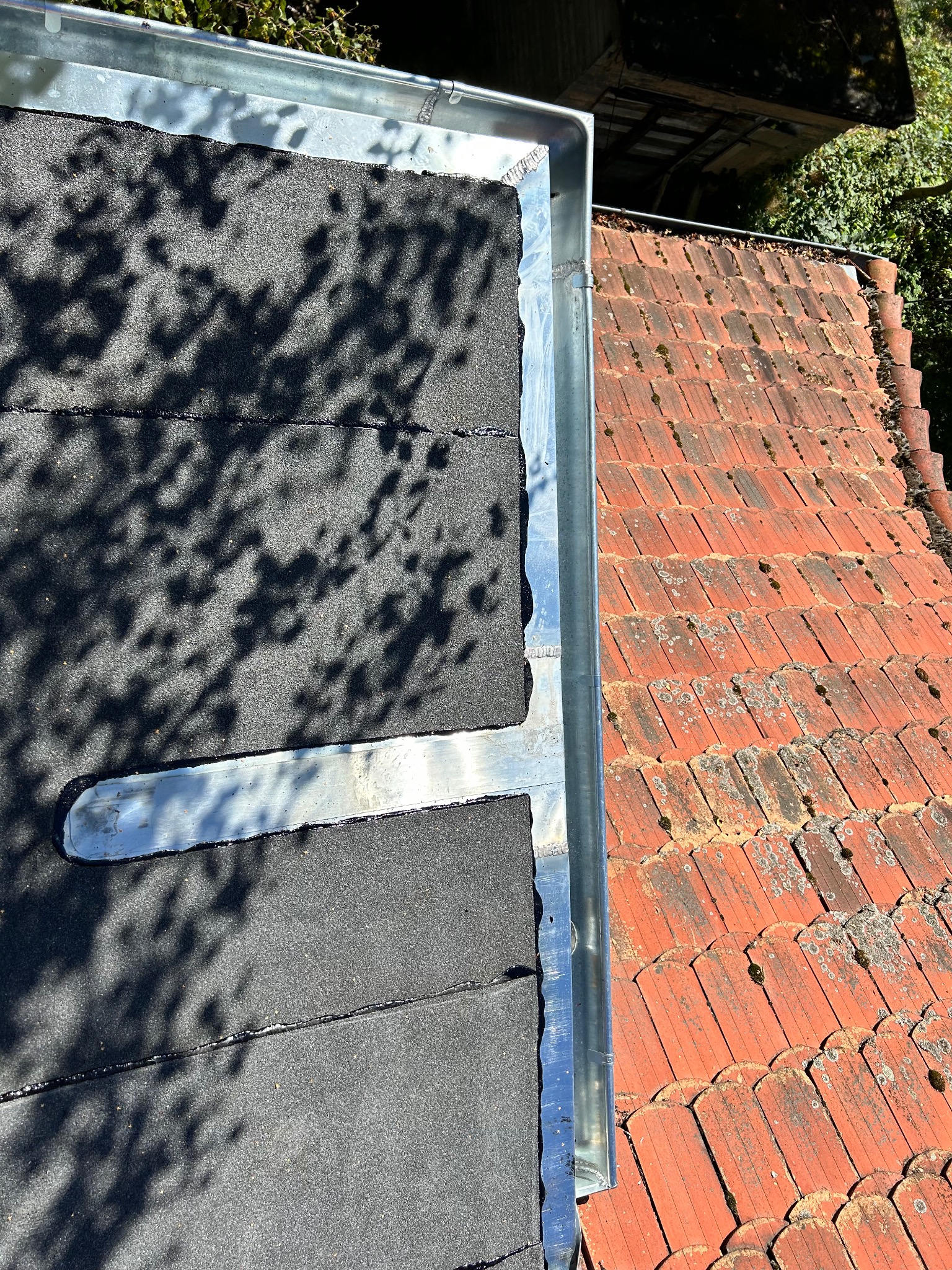 Widok z góry na fragment dachu z papy z obróbką blacharską oraz fragment dachu krytego czerwoną dachówką karpiówką, z widocznymi śladami mchu i zabrudzeń.